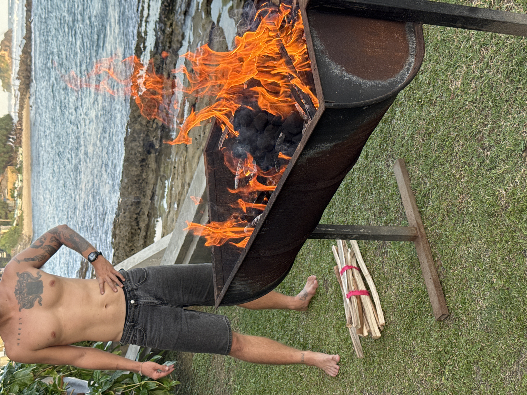 A shirtless man with tattoos on his torso and arms stands on a grassy area near the ocean in Goviyapana, Sri Lanka. He is wearing denim shorts and a watch on his left wrist. To his right, a metal barbecue grill is filled with burning charcoal and wood, emitting large flames. A pile of dry sticks tied with pink ribbon lies on the grass to the left of the grill. In the background, the ocean waves crash against rocky shores, and a small island with vegetation is visible in the distance. The overall scene suggests an outdoor cooking or gathering event.
