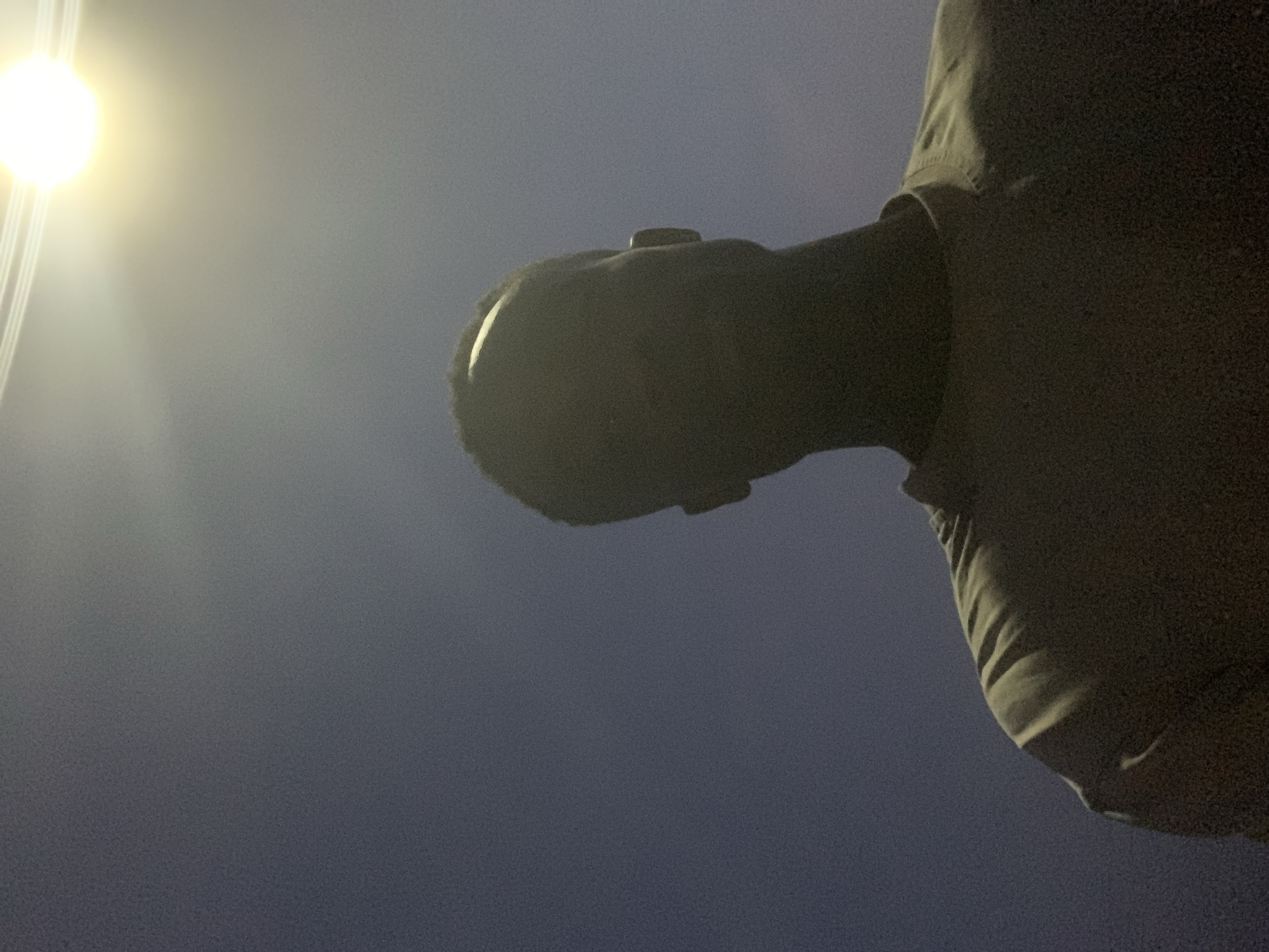 A low-angle shot in Gbeleko, Nigeria, shows a person's head and shoulder against a dark sky, with a bright street light to the left. The person appears to be wearing a dark-colored shirt. The person is standing beneath a street light in the dark, looking up at the light.