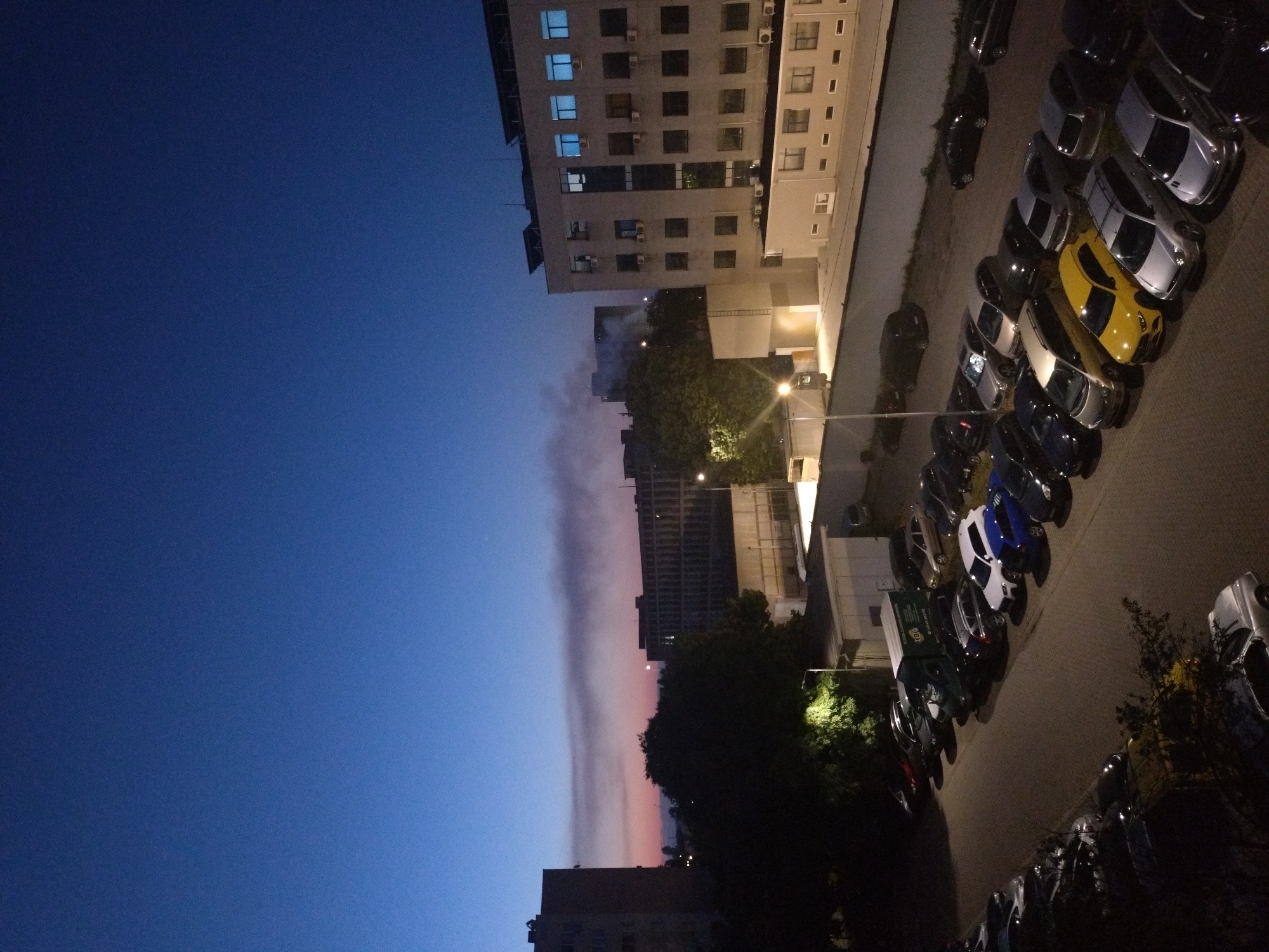 The image depicts a view of Stara Zagora, Bulgaria, at dusk or dawn. The sky is a gradient from deep blue to lighter hues with a distinct, vertical cloud formation tinged with pink. Buildings of varying heights are visible; one is a light-colored apartment building. A line of parked cars occupies the right side of the frame. The scene is illuminated by streetlights and some ambient light.