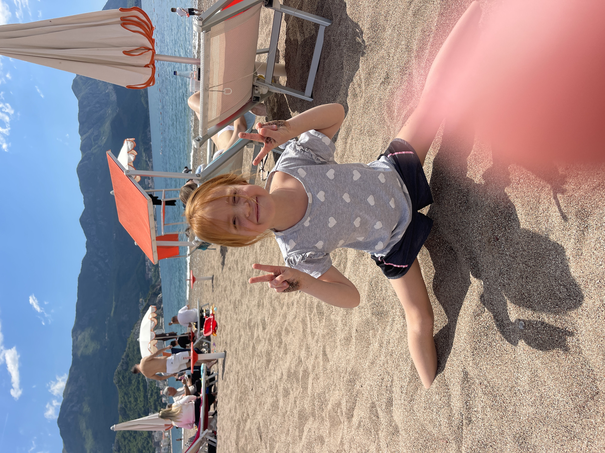 The image shows a young girl kneeling on a sandy beach. She has red hair and is wearing a gray t-shirt with white hearts and dark shorts. Her hands are covered in sand, and she is making peace signs with both of them. The girl is smiling at the camera. In the background, there is a beach with sun loungers and umbrellas, as well as a body of water and a large mountain. People are visible in the background. The overall scene suggests a sunny day at the beach in Boreti, Montenegro.