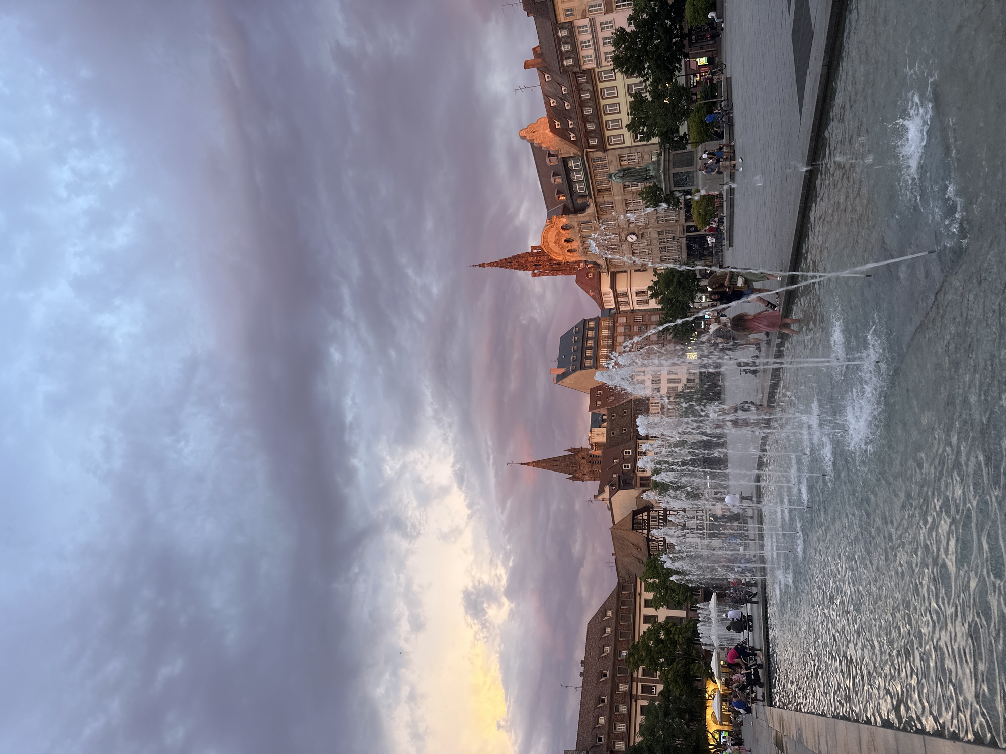 The image shows a fountain in the Place de la Cathédrale in Strasbourg, France, at dusk. The sky is filled with dramatic, cloudy hues of purple, pink, and orange. In the background, the iconic Strasbourg Cathedral and other historic buildings with traditional Alsace architecture line the square. People are visible in the distance, some sitting on benches and others walking. Children are playing in the fountain. The fountain itself features multiple jets of water creating a dynamic and refreshing scene in the urban setting.