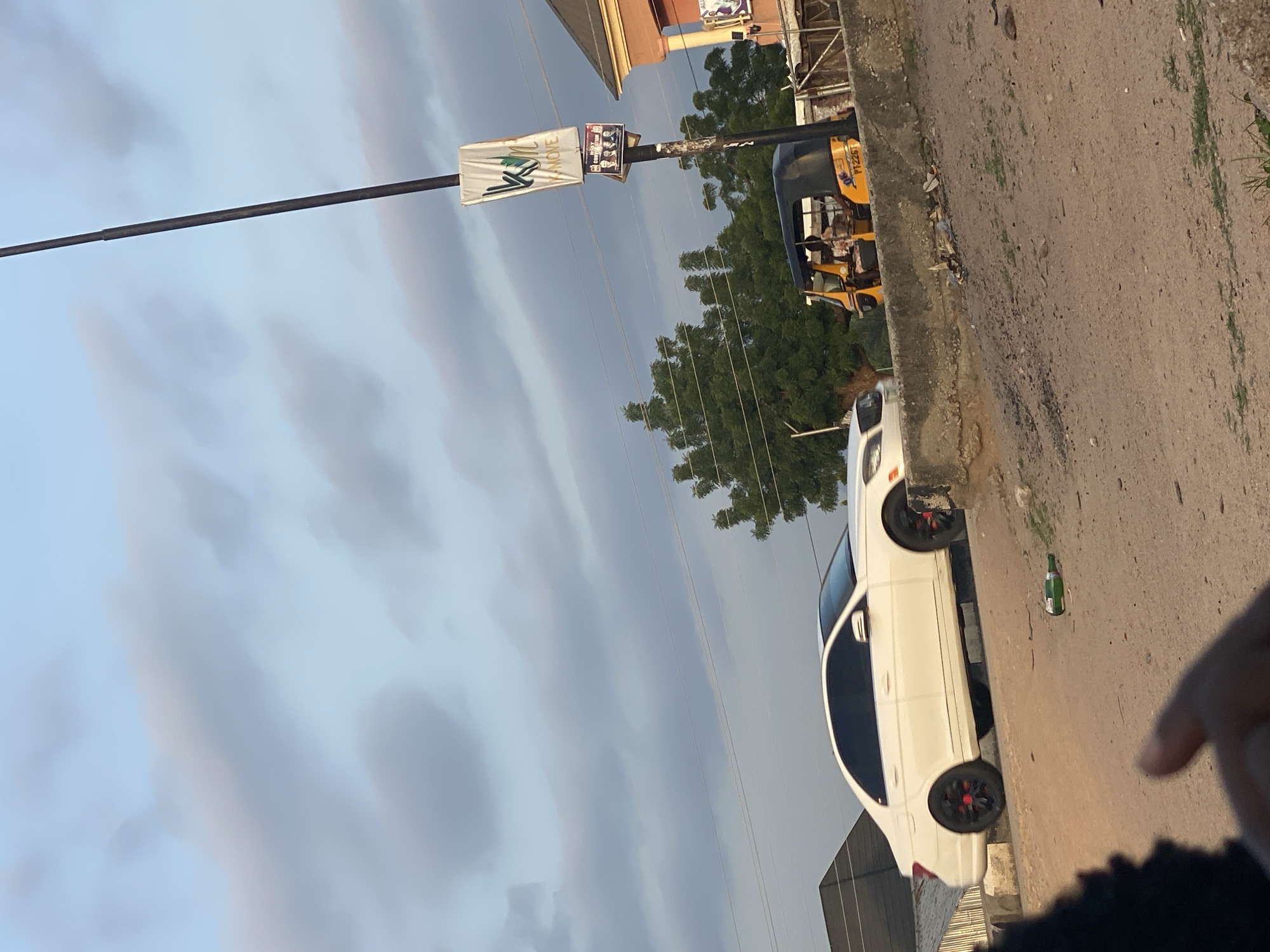 The image depicts a street scene in Jalingo, Nigeria, under a cloudy sky. In the foreground, a sandy, uneven ground is visible, with a green bottle lying on its side. A blurred hand and arm are in the lower left corner, suggesting a first-person perspective. A white Mercedes-Benz sedan with red accents on its wheels is parked on an elevated concrete barrier. To the right, a yellow auto-rickshaw, a common mode of transport in the region, is also on the elevated barrier, with passengers inside. A tall lamppost stands prominently, adorned with two banners. The upper banner is white with a green logo and text reading "KM MOVE," while the lower banner displays photos of four men. Behind the vehicles and lamppost, lush green trees form a backdrop, and a building with an orange facade is partially visible on the far right.