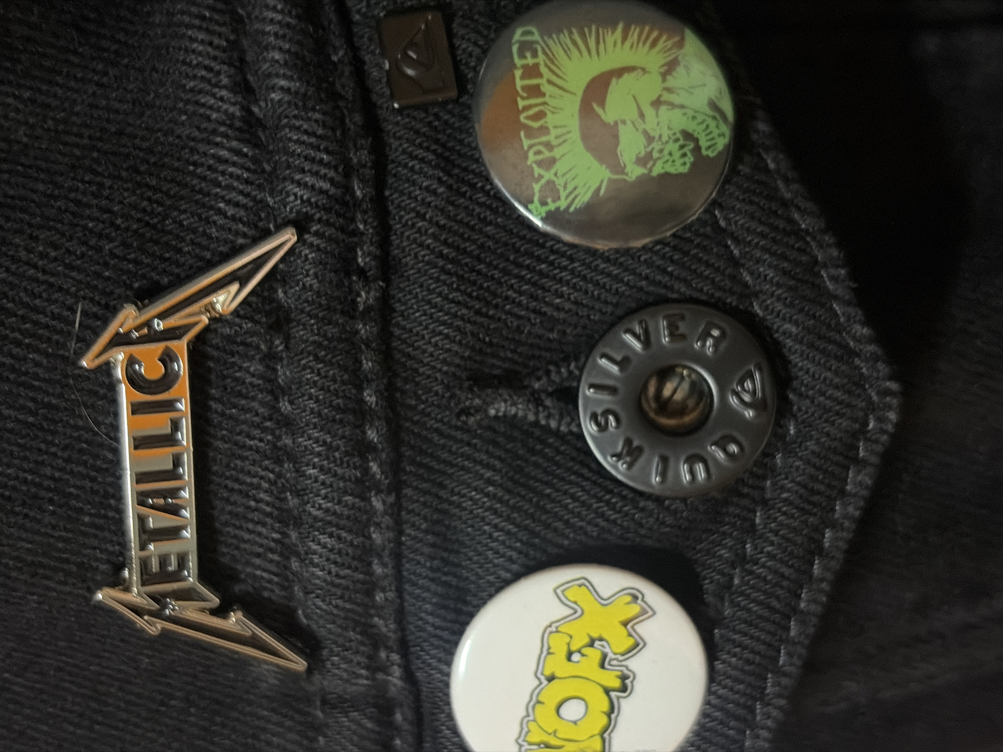 The image depicts a close-up of a black denim jacket. Several pins are attached to the jacket. From top to bottom: a black pin with a green image of a punk figure and the word "EXPLOITED", a square pin, a black button with the inscription "SILVER Quik", a silver Metallica logo pin, and a white pin with yellow lettering. The location and city are not discernable from the image. There are no people visible.