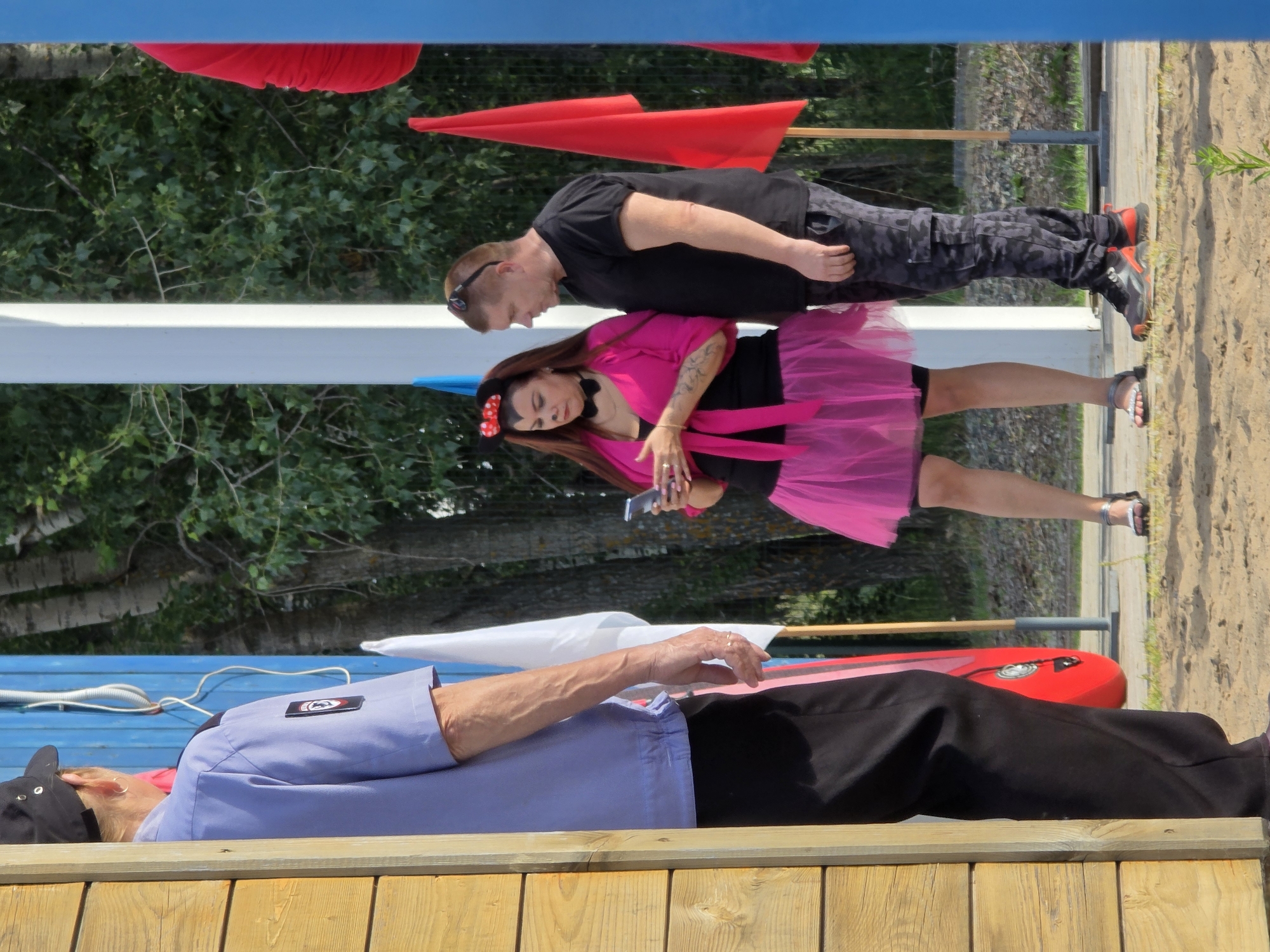 The image depicts an outdoor scene with three individuals. In the foreground, lying on a wooden structure, is a person wearing a blue shirt and black pants, with a black cap on their head. To their right, a red object resembling a kayak or canoe is visible.
In the background, two people are interacting. A man, wearing a black t-shirt and camouflage pants, is leaning over a woman who is wearing a pink dress. The woman is holding a smartphone. Behind them, there are trees and a sandy area. Red fabric is draped in the upper portion of the image, and blue surfaces are also visible. The scene appears to be set during daytime with clear weather. The overall impression is a casual outdoor gathering or event. There is a visible logo on the blue shirt of the person in the foreground, but it is not legible.