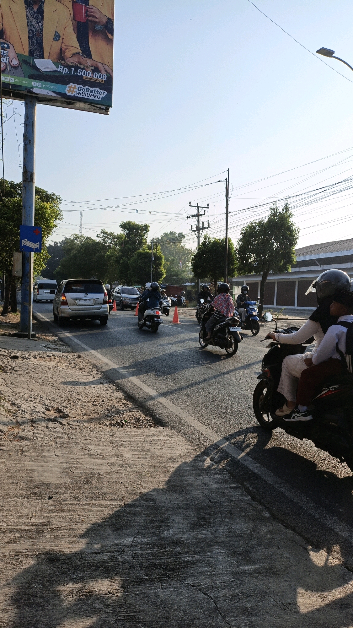 The image depicts a street scene in Kuwasen, Indonesia, on a clear, sunny day with a bright blue sky. In the foreground, a concrete sidewalk with visible cracks is shaded by the long shadow of a motorcycle and its riders. The asphalt road shows lane markings and is populated with several motorcycles.
Several individuals are riding motorcycles. On the right side of the frame, a motorcycle carries two passengers; the rider wears a black helmet and a black hijab, with a patterned scarf visible. The passenger behind wears a white shirt, maroon pants, and a black backpack. Further down the road, another motorcycle is ridden by a person in a black helmet and a dark top, with a patterned shirt visible underneath. A blue motorcycle is also visible with a rider. In the middle ground, two more motorcycles are seen, one with an orange traffic cone behind it, and another with a rider wearing a dark helmet and a red and white checkered shirt.
Several cars are also present. A silver SUV is stopped on the left side of the road, preceded by a white van. Another car, possibly dark grey, is behind the silver SUV.
The background features a billboard on the left side of the image, with text and images related to an educational institution. Trees line the sides of the road, with some foliage obscuring buildings in the distance. Electrical poles and wires are visible against the sky. Orange traffic cones are placed on the road, suggesting a temporary lane closure or traffic management. The overall impression is a typical day in a suburban or semi-urban Indonesian environment with moderate traffic.