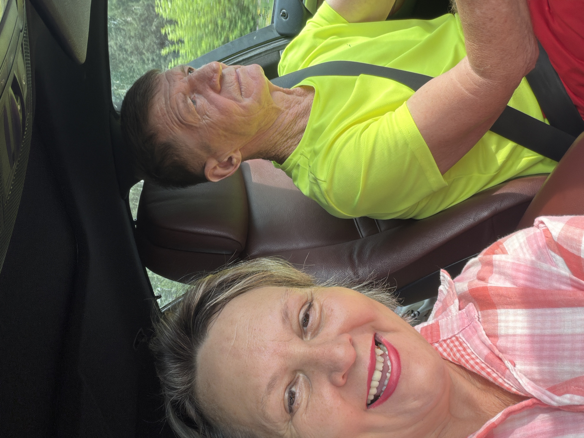 A selfie is taken from the passenger seat of a car, showing two individuals inside. The woman in the foreground, on the left, smiles broadly at the camera, her makeup including bright red lipstick. She wears a pink and white gingham shirt. Behind her, on the right, a man is driving. He looks to his right, away from the camera, and wears a bright neon yellow t-shirt and red shorts. A black seatbelt is visible across his chest. The car's interior, including brown leather seats and a black headliner, is visible. Outside the car windows, lush green trees and foliage are visible, suggesting a rural or suburban setting, possibly during daylight hours. The scene is captured in a mobile phone selfie.
