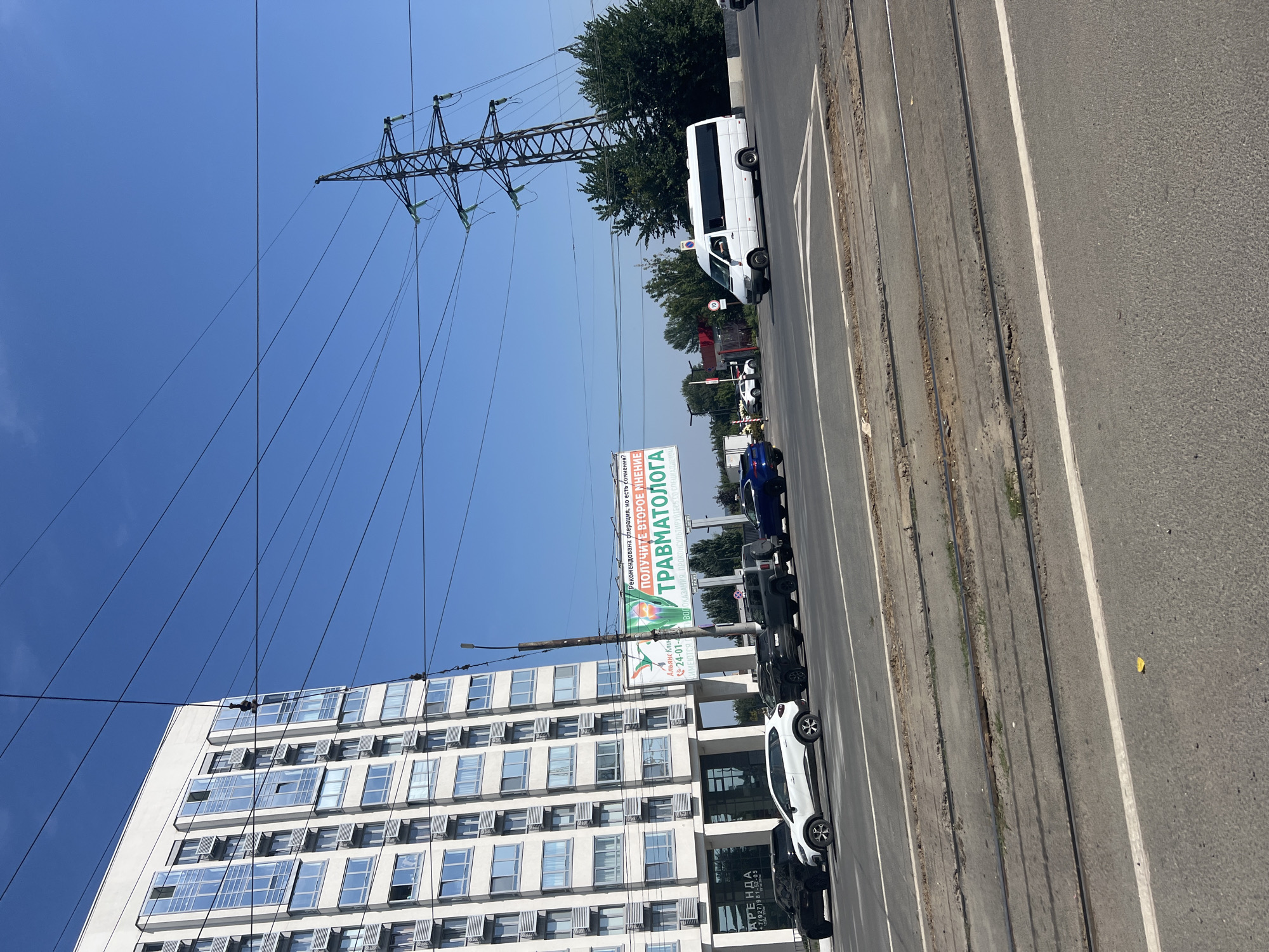 The image captures a street scene in Ulyanovsk, Russia, under a clear blue sky. In the foreground, a road with visible tram tracks is present. Several cars are driving on the road, including a white minivan, a dark blue SUV, and several other vehicles. On the left side of the image, a modern, multi-story building with a white facade and many windows is visible. To the right of the building, a large billboard with text and graphics is displayed, promoting a trauma center. In the background, a tall electricity pylon with numerous wires stretches across the sky, and lush green trees are scattered throughout. The overall environment suggests a daytime urban setting.