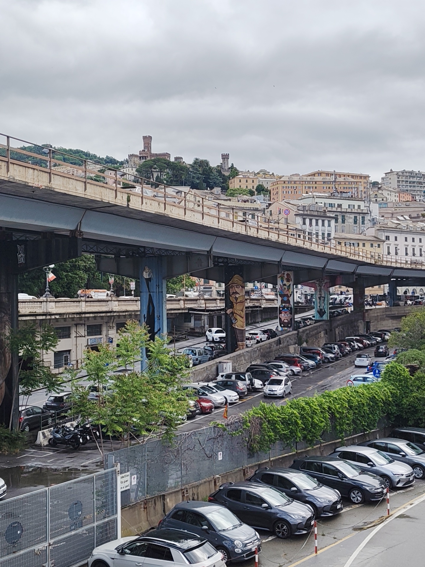 The image shows an elevated urban scene with multiple layers of infrastructure and buildings, likely in Genoa, Italy, given the architecture and overall urban arrangement. A large, gray bridge or elevated road structure dominates the middle ground, with artwork painted on the supporting pillars. Below the bridge are rows of parked cars on various streets, and the presence of trees and vegetation hints at green spaces among the urban development. In the background, residential and commercial buildings are visible on the hills, with a medieval-looking castle complex at the top. The sky is overcast, suggesting a cloudy day.