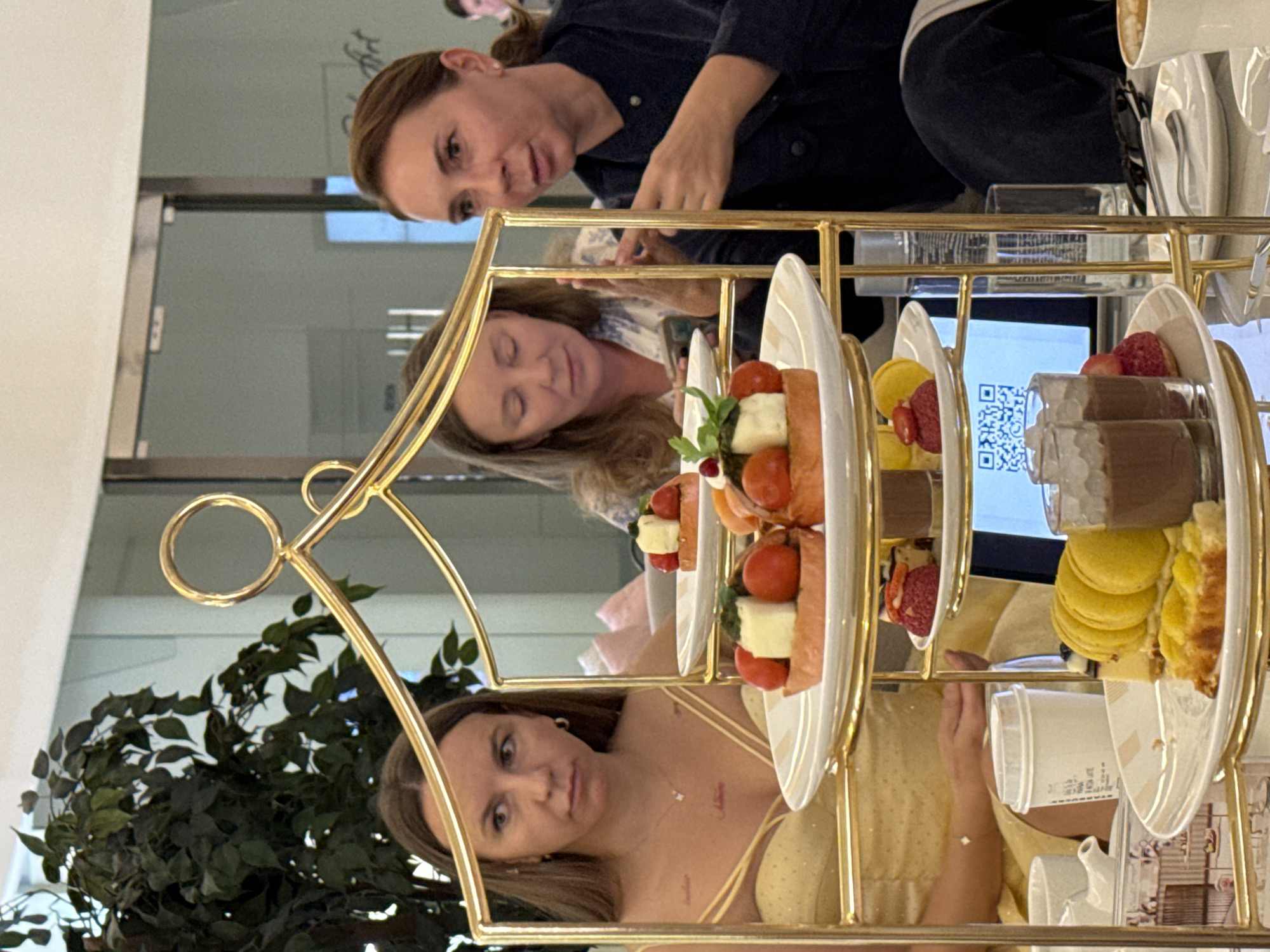 The image, oriented 90 degrees clockwise, captures an indoor gathering in what appears to be a cafe or restaurant. Three women are visible around a table featuring a prominent gold-colored tiered food stand.
In the upper part of the frame, a woman with light brown hair, wearing a dark blue top, looks towards the camera with a neutral expression. Below her, reflected in a glossy surface, is another woman with long light brown hair, wearing a white and blue patterned top, seemingly occupied with a smartphone. In the lower portion of the frame, a third woman with long light brown hair, dressed in a light yellow strapped dress, also looks towards the camera with a calm demeanor. A partial reflection of another person's head is faintly visible in the upper right.
The central tiered stand holds a variety of refreshments. The middle tier displays savory items, likely mini sandwiches or tartlets, topped with cherry tomatoes and white cheese, garnished with herbs. Also on this tier are yellow macarons and small glasses containing chocolate mousse topped with whipped cream, alongside other small pastries with red berries. The bottom tier includes more yellow macarons and pastries. A white ceramic teacup or coffee cup and a white disposable cup, clearly showing the "STARBUCKS" logo, are also present.
A clear glass water bottle is visible on the table behind the food stand. In the background, a reflection on a surface reveals a blue screen displaying a white QR code. A large potted plant with green foliage is on the left. The scene suggests a relaxed afternoon tea or dessert occasion. Faint, stylized text can be seen on a mirrored wall in the reflections. The gathering is set indoors, possibly in Dubai, United Arab Emirates, as per the provided location context.