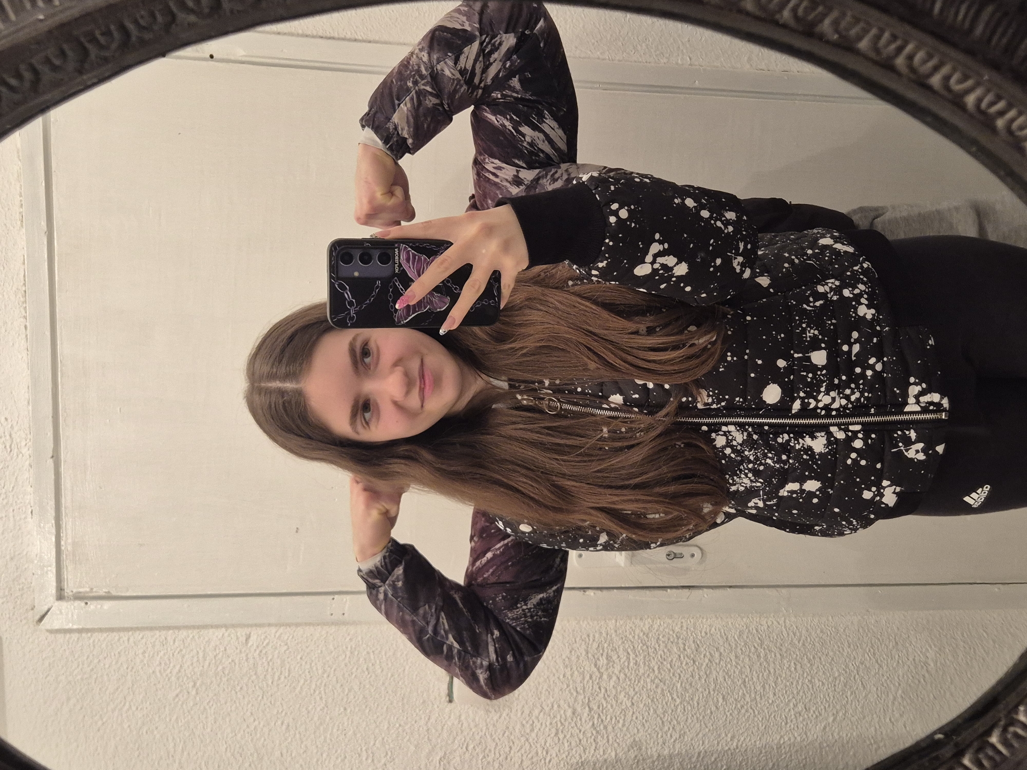 A young woman is taking a selfie in a mirror. She is wearing a black jacket with white splatters, black pants, and has long brown hair. She is holding a cell phone with a black case with a butterfly design. The background appears to be a plain, white wall. The mirror has an ornate, dark frame. The photo may be taken indoors. There is no information suggesting a location in the city of Pavlikeni, Bulgaria.