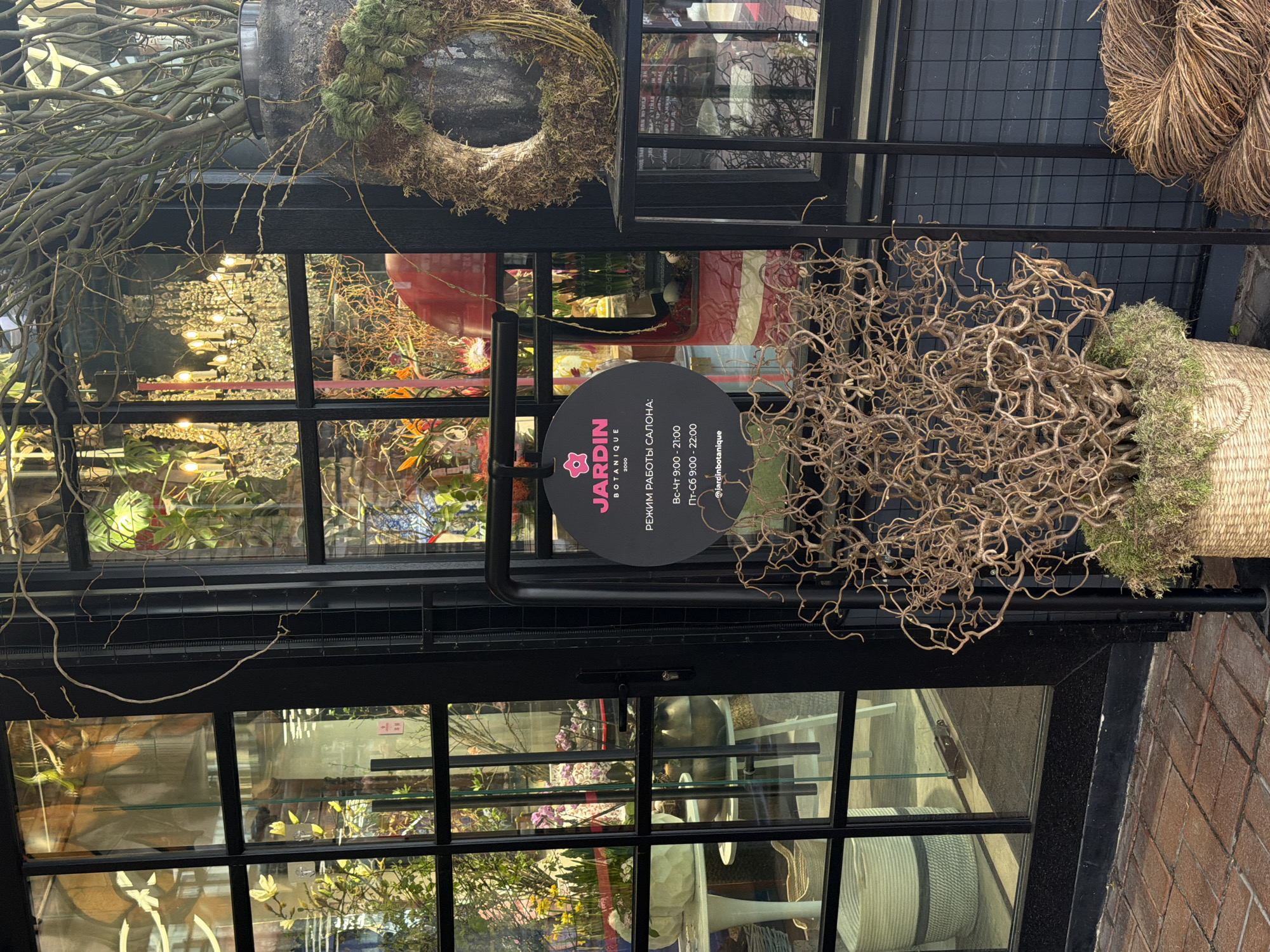 The image shows the exterior of a shop named "JARDIN BOTANIQUE" located in Moscow, Russia. The shop's hours are displayed on a round, black sign written in Russian. The sign indicates that the shop is open from 9:00 to 21:00 from Sunday to Thursday, and from 9:00 to 22:00 on Friday and Saturday. The shop's Instagram handle "@jardinbotanique" is also visible on the sign.
The shop's facade includes a black metal grid frame that creates multiple window panes. The interior of the shop can be seen through the windows, revealing various floral arrangements, plants, and decorative items. A wreath, as well as some dried branches and moss, are attached to the exterior of the shop. The shop appears to be located on a brick sidewalk.