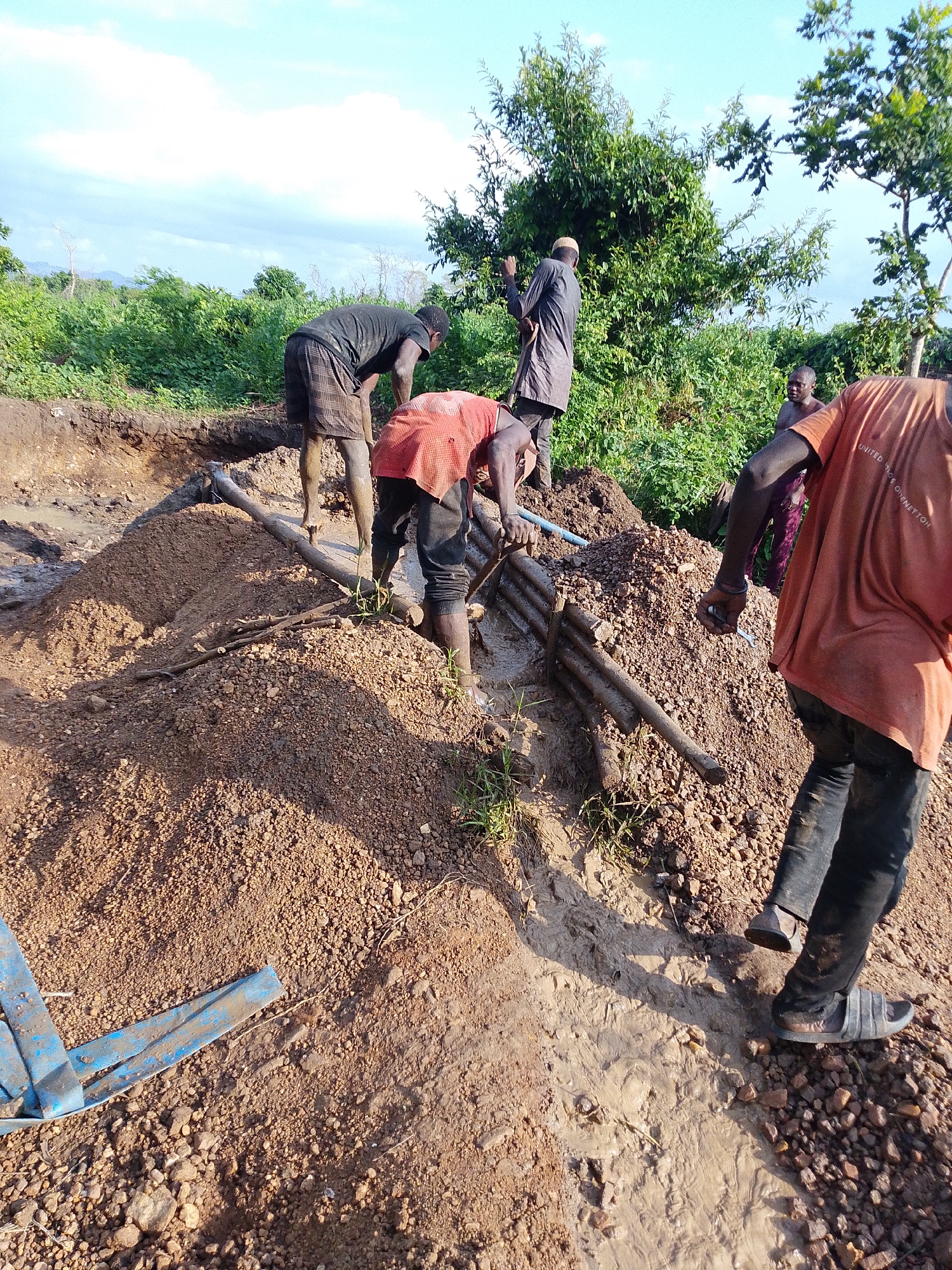 In Otulu, Nigeria, a group of men are engaged in manual labor at a construction or excavation site. The scene is muddy with piles of earth and debris. Several individuals are actively working, some appearing to be digging or moving soil. One man in an orange shirt is positioned in the foreground, facing away from the camera and appearing to be working with a pipe or conduit. Another man in a black shirt and checkered shorts is bending down, seemingly involved in digging. A third individual, dressed in a dark robe and cap, stands near a pile of earth, holding a shovel. The environment is outdoors, with lush green vegetation and trees visible in the background, under a bright sky with some clouds. The ground is a mix of soil, mud, and small rocks. There is a blue plastic object visible in the lower left corner of the frame, partially submerged in mud. The overall impression is one of hard physical work in a rural or undeveloped setting.