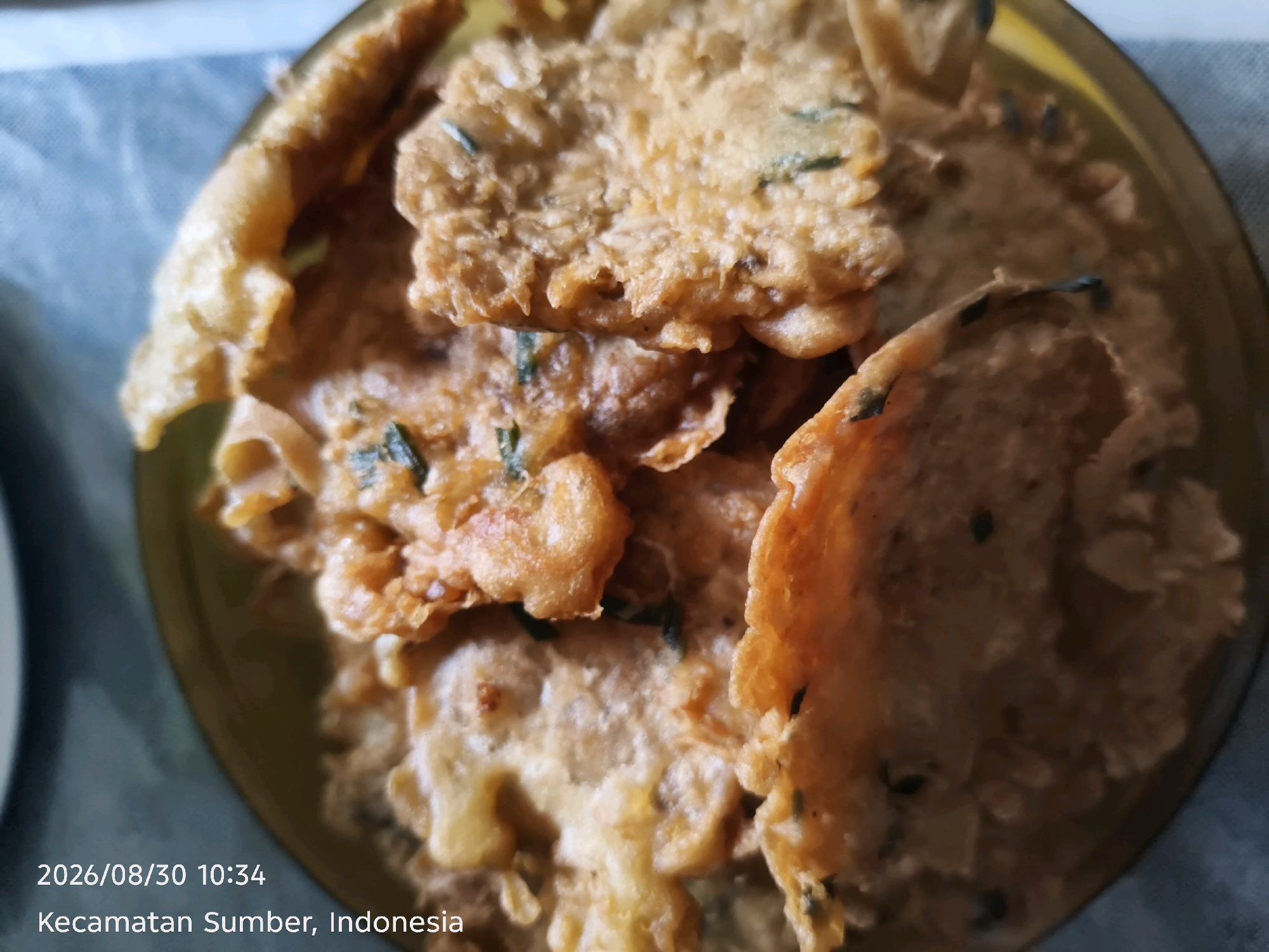 A close-up, overhead shot shows a shallow bowl filled with fried Indonesian tempeh snacks. The tempeh pieces are irregular in shape and golden brown, with visible green herbs mixed into the batter. The bowl itself appears to be a dark yellow or brown color and rests on a blue and white patterned cloth. The bottom of the image contains a timestamp and location: "2026/08/30 10:34 Kecamatan Sumber, Indonesia."