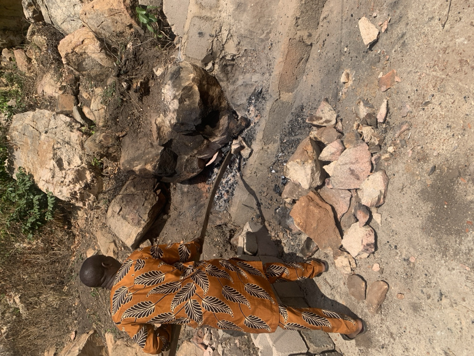 In Egbedi, Nigeria, an adult male stands with his back to the viewer, facing a functional fire pit or forge constructed amidst natural rock formations and concrete blocks. He is dressed in a full-length, two-piece orange-brown garment featuring a black and white leaf pattern, paired with light brown sandals. The man uses a long, dark metal rod to manipulate glowing embers and a visible reddish-orange object within the active fire. The immediate ground is composed of dry, light-colored soil, with a cluster of various sized rocks and broken masonry fragments in the foreground. The background features large, weathered rocks and sparse green vegetation under direct sunlight.