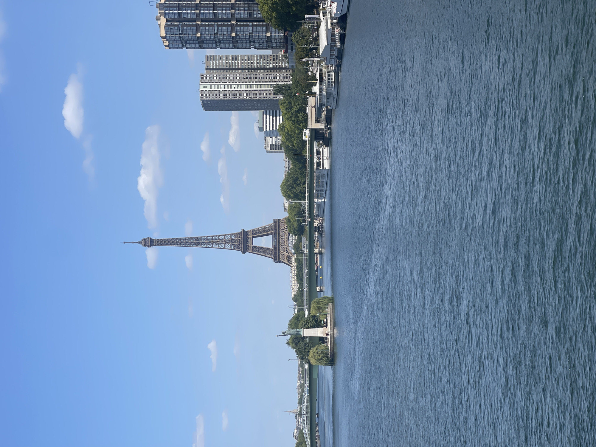 This image shows a view of the Seine River in Paris, France, with the Eiffel Tower prominently visible in the background. On the left side of the image, a small island in the river features a replica of the Statue of Liberty, surrounded by trees. A bridge spans the river in the midground, with various boats docked along the riverbank on the right side. Several modern apartment buildings rise behind the riverfront. The sky is blue with scattered white clouds. The water is a deep blue with small ripples. There are no people clearly visible in the foreground or midground, but there may be individuals on the island or near the docked boats. The scene depicts a sunny day with a clear sky.
