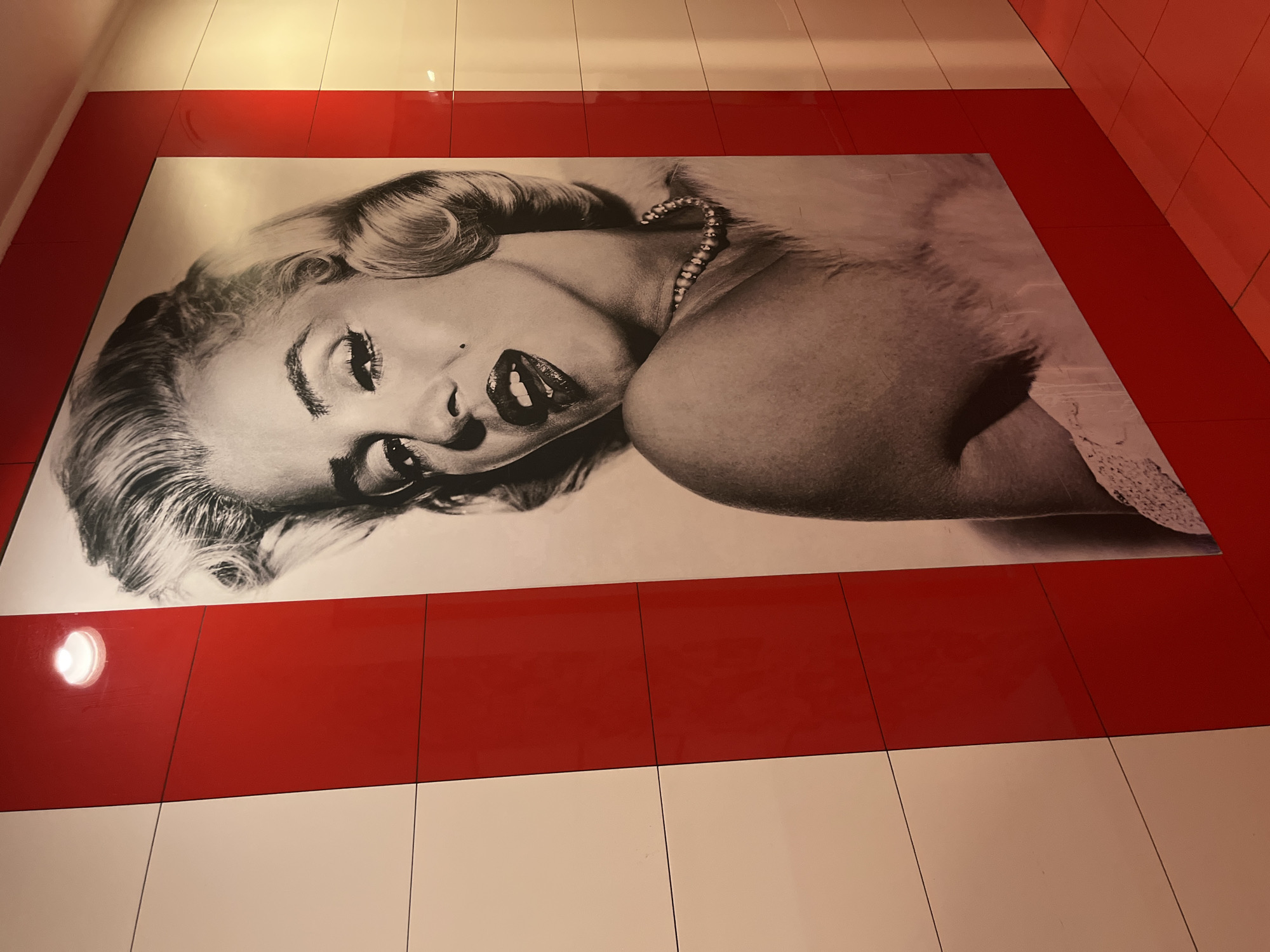 This interior scene in Sochi, Russia, features a large, monochrome photographic portrait of Marilyn Monroe displayed on a wall. The portrait captures Monroe from the upper chest, gazing forward with a partially open mouth, a beauty mark above her lip, and her hair styled in characteristic waves. She is adorned with a pearl necklace and what appears to be a light-colored fur or feather boa over her left shoulder. The wall around the portrait is composed of alternating glossy red and matte white rectangular panels, arranged vertically to the left and as framing elements around the image. Horizontal seams delineate individual panels. A distinct circular light source is visibly reflected on an upper-left red panel. The floor is covered with glossy, square red ceramic tiles, which reflect ambient light. The overall environment is a brightly lit, stylized indoor space, likely a commercial or public area.