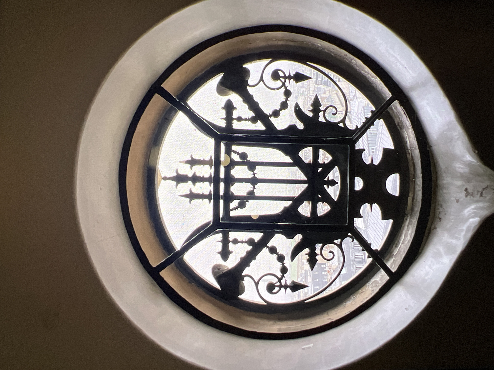 This image shows a close-up view of a round, ornate window with dark metalwork. The window is framed by a thick, light-colored material, possibly plaster or stone, which has a slightly rough texture. The metalwork inside the window is intricate, featuring decorative elements that resemble finials, arrows, and beaded strings. These elements divide the window into several sections, through which a bright, hazy scene of buildings can be seen. The architectural style of the buildings in the background suggests an urban setting, possibly with historical significance. The lighting is bright and diffused, indicating it might be daytime. There is no visible text in the image. The overall impression is one of looking through a decorative grate from an interior space out to a city view. The context of Verona, Italy, suggests this could be a historical building or landmark.