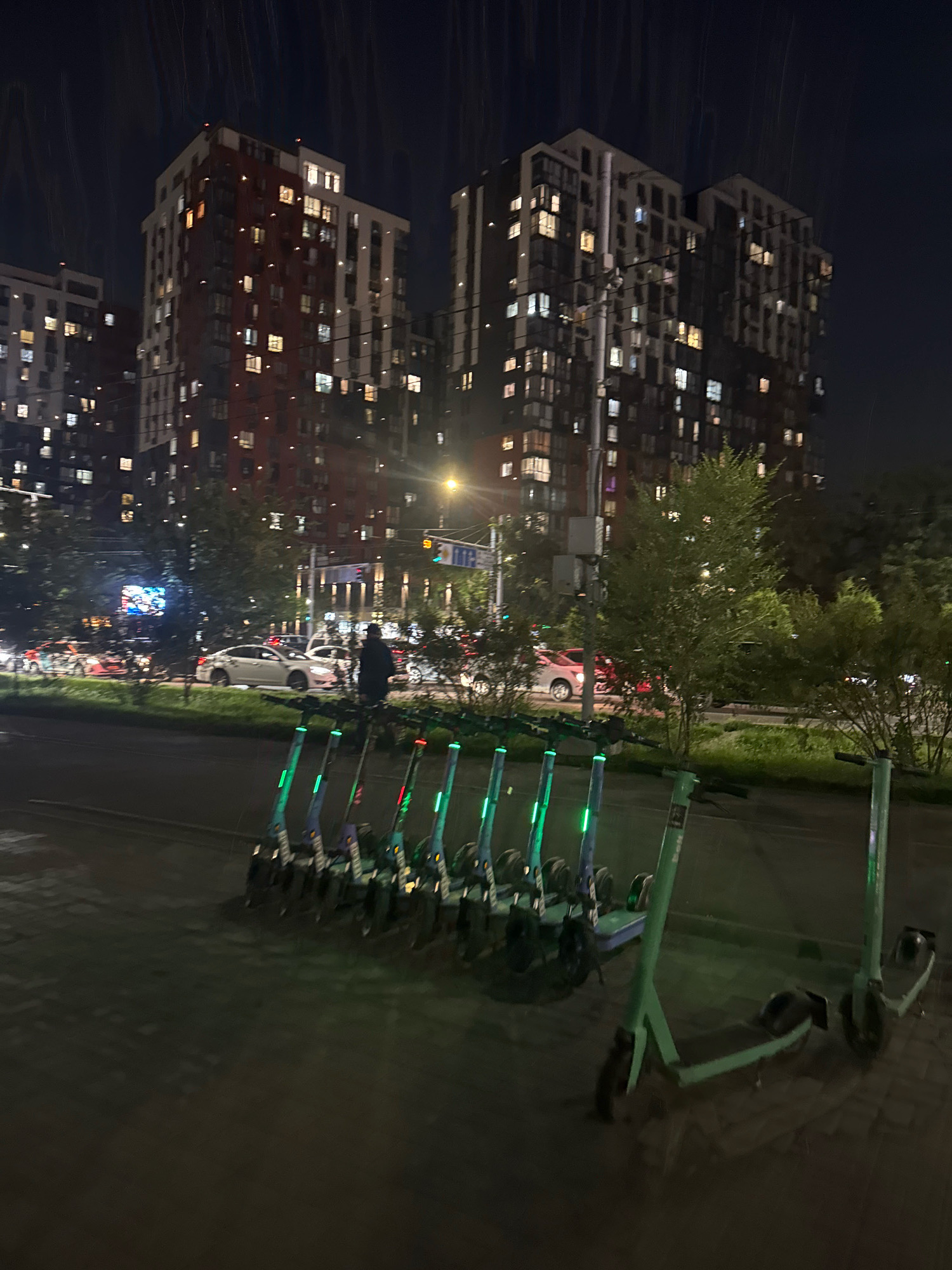 This image depicts a street scene at night in Almaty, Kazakhstan. In the foreground, a row of electric scooters with illuminated green accents is parked on a paved surface. Some of the scooters are fully visible, while others are partially obscured. Beyond the scooters, a street runs parallel to the parking area, with cars moving in both directions. The cars are illuminated by their headlights and taillights, creating streaks of light due to motion blur.
In the background, several multi-story apartment buildings are visible. These buildings are illuminated from within, with many windows showing lights. Trees and streetlights are also present, adding to the urban nighttime ambiance. The sky is dark, indicating that it is nighttime. The overall impression is of a busy, modern city at night, with various modes of transportation and residential areas coexisting.