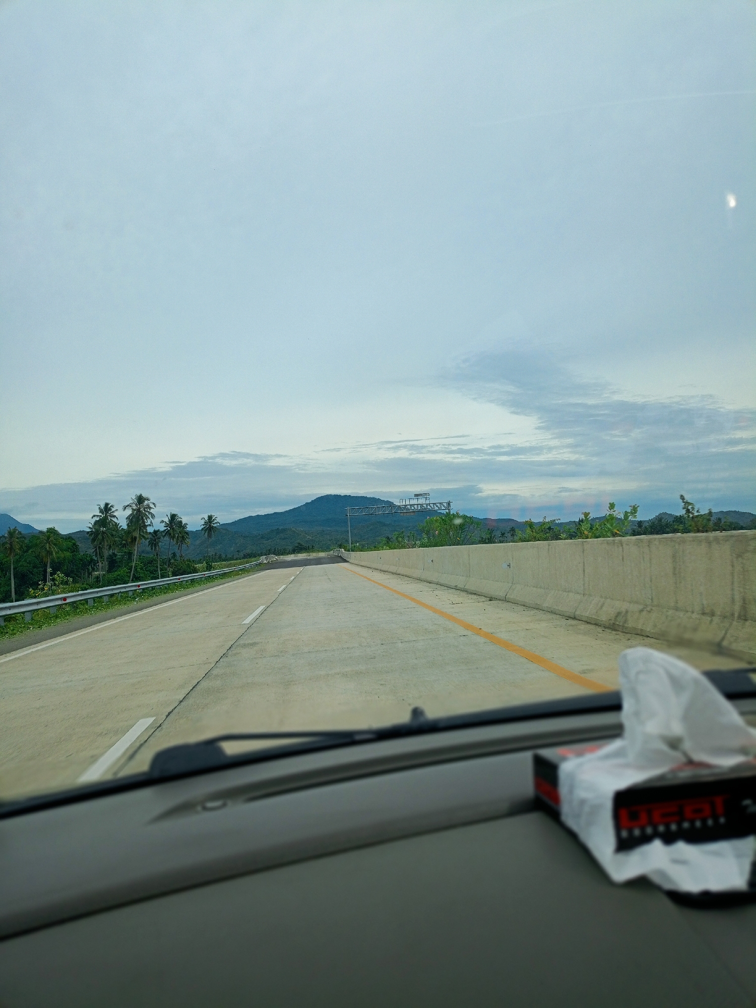 Captured from the dashboard perspective of a vehicle in motion, the scene presents a clear asphalt road cutting through a landscape in Suyo Paloh, Indonesia. The road is marked with a yellow line down the center and a white line on the left side, with concrete barriers lining the right edge of the road. The backdrop features green vegetation, palm trees, and rolling hills under a partly cloudy sky. A tissue box is visible on the dashboard.