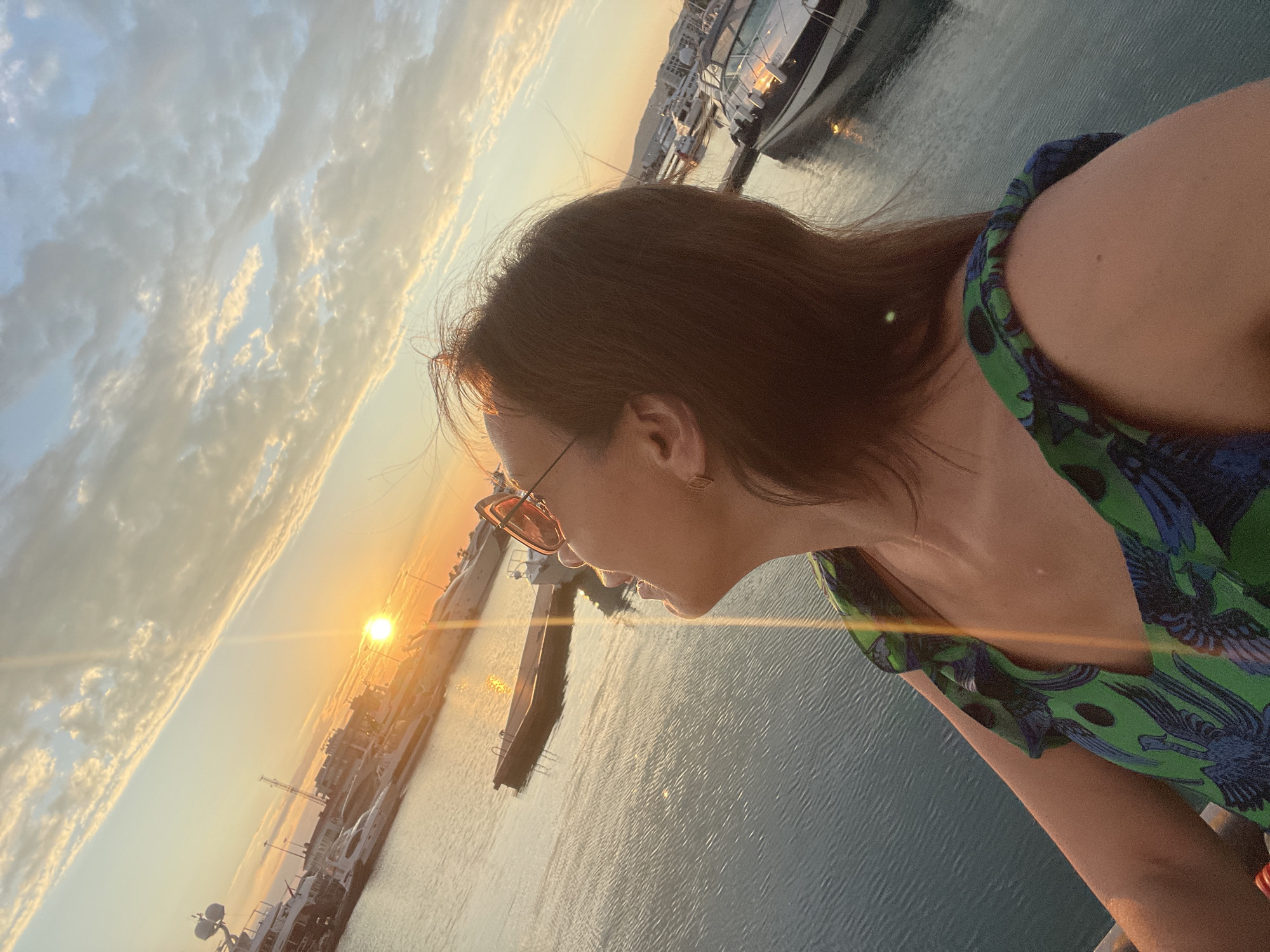The image shows a young woman taking a selfie at sunset in a harbor. She is wearing a green and blue patterned dress and glasses. The sun is setting over the water, casting a warm glow on the scene. Boats are docked in the harbor, and the sky is filled with clouds. The woman appears to be relaxed and happy, enjoying the beautiful view. The location is Sochi, Russia.