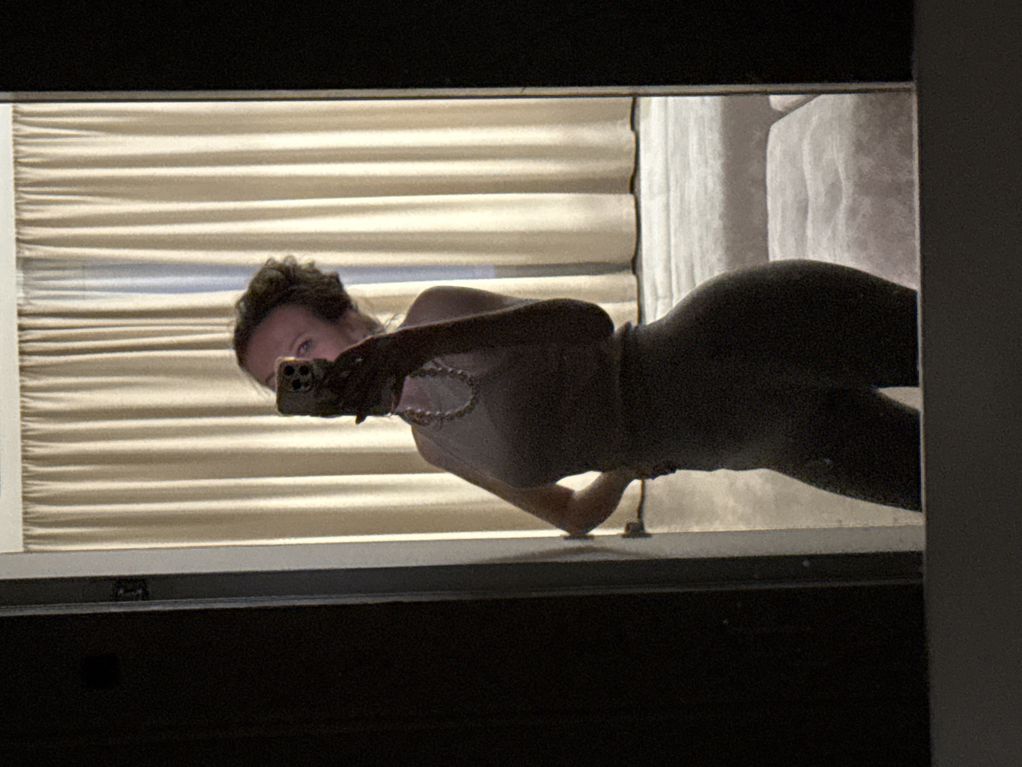 The image is a mirror selfie showing a person taking a photograph with a smartphone. The subject, likely a woman, has short, dark, curly hair and is wearing a light-colored top (possibly a camisole or tank top) and dark-colored pants or leggings. A necklace is visible around their neck. They are holding the smartphone with their right hand, which is extended, and the phone partially obscures their face, with one eye clearly visible looking towards the camera.
The background visible in the mirror's reflection consists primarily of light beige or cream-colored horizontal pleated blinds or curtains, suggesting a window behind the person. To the right of the person, a segment of a white or light-colored bed or sofa with bedding or upholstery is visible. The lighting is indoor, appearing somewhat dim or controlled, possibly indicating evening time or a room with closed blinds. The setting appears to be an indoor space, likely a bedroom or living room. The location context provided is Khimki, Russia. There is no visible text in the image.
