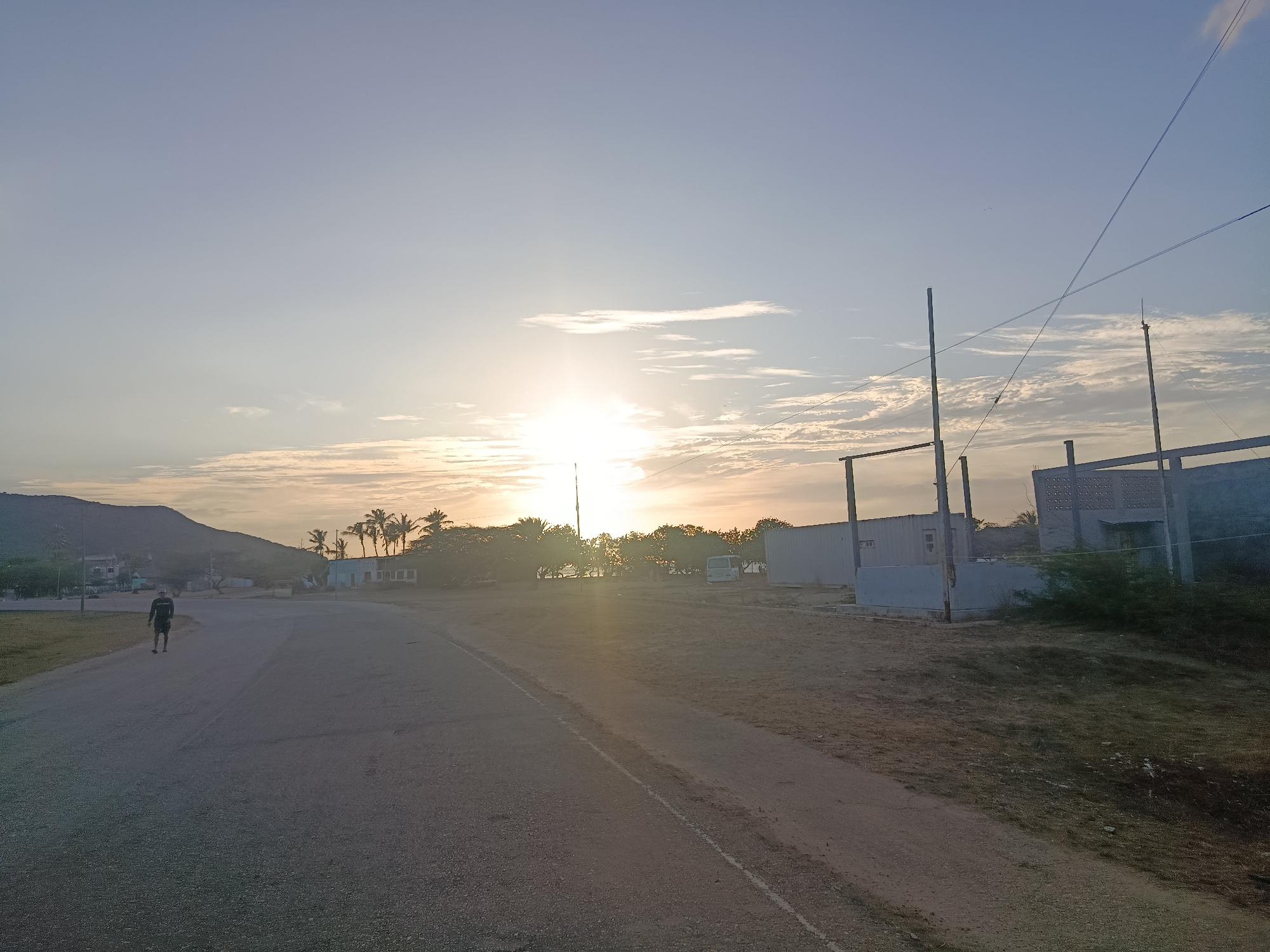 The image captures a scene at dusk or dawn in what appears to be a coastal area near Altagracia, Venezuela. The sun is partially obscured by clouds and trees on the horizon, casting a bright glow. In the foreground, a paved road stretches away from the viewer, transitioning into a dirt shoulder on the right. A single individual is walking on the left side of the road, away from the camera. The environment includes a small mountain on the left, trees (some palm trees), and some buildings and structures along the right side of the road, including shipping containers and an unfinished or partially constructed building. Overhead power lines are visible in the sky. The lighting suggests a time of day with low, angled sunlight.