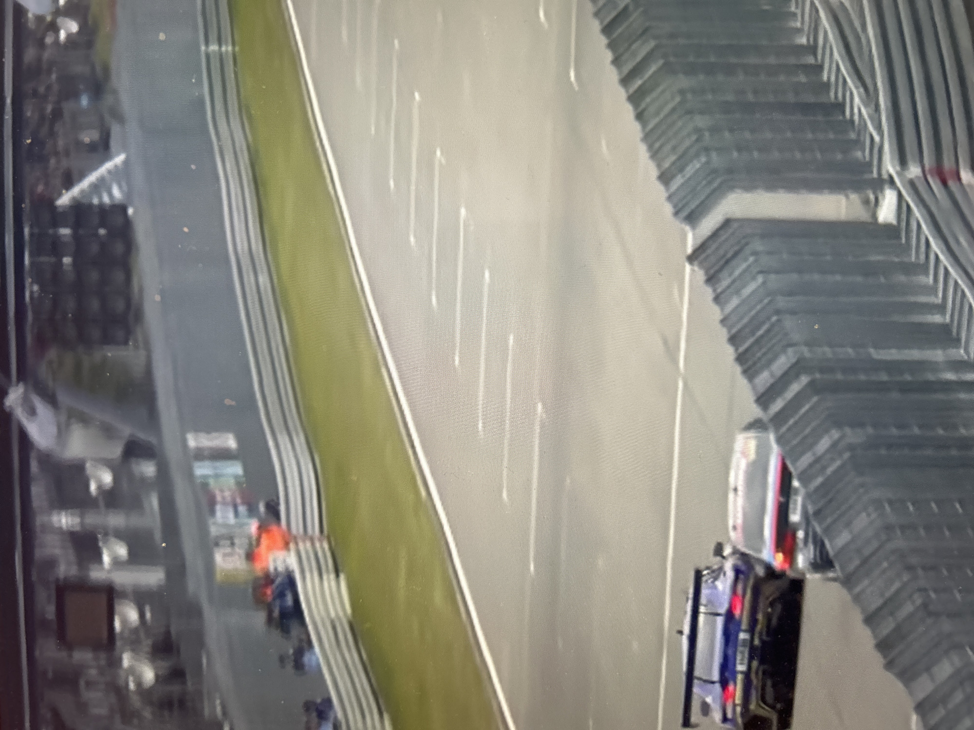 The image shows an aerial view of a racetrack. A grassy median separates the track from a pit lane, with cars and personnel visible. White lines mark the racing surface. To the right, a grandstand structure is visible. The location is not identifiable as Sofia, Bulgaria, based on the image content.