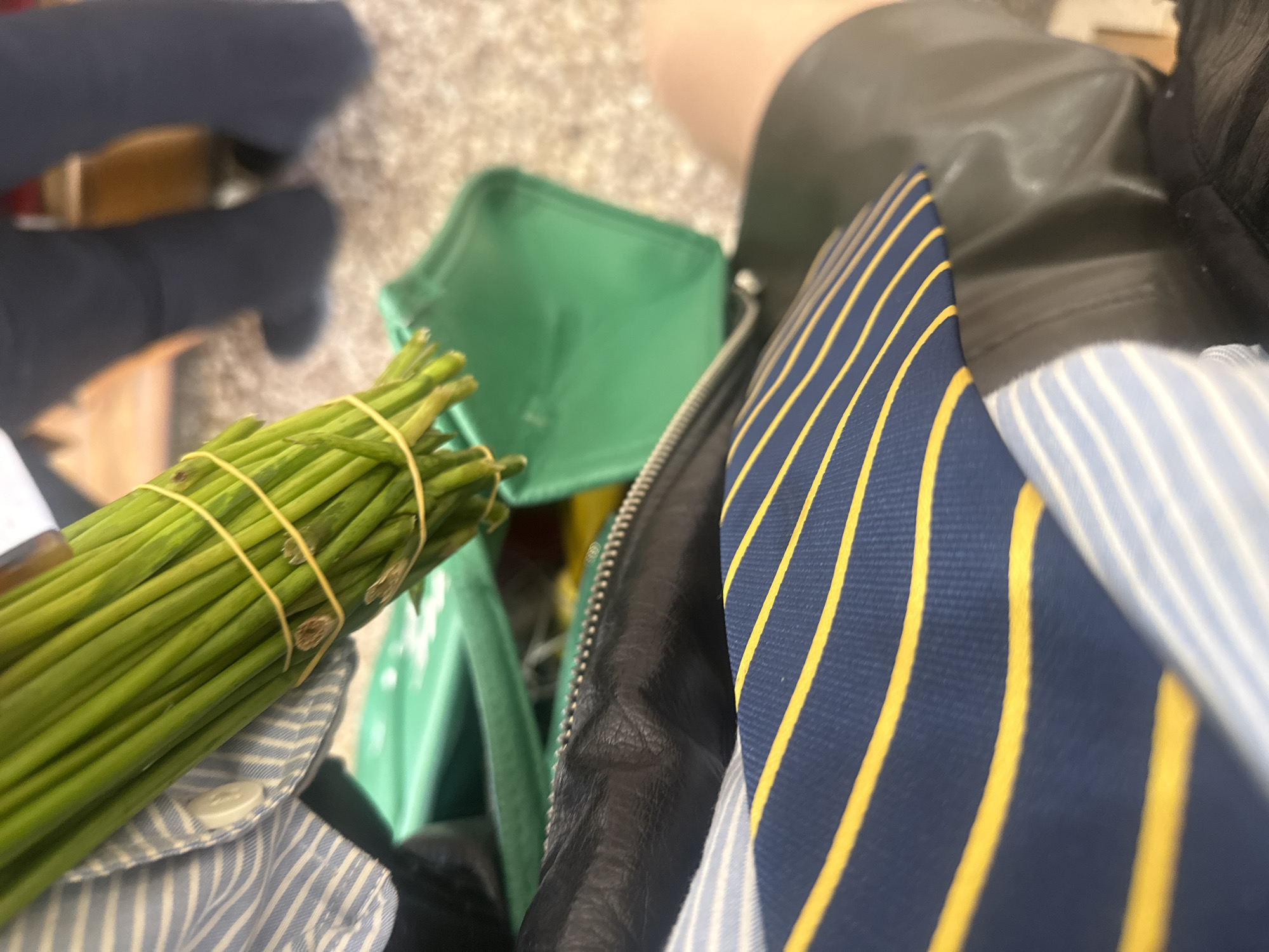 A close-up shot shows a bundle of green chives secured with two rubber bands, resting atop a light blue and white striped button-down shirt. Beside the chives, there's a green bag. Adjacent to the bag and shirt is a black leather jacket with a visible zipper. A dark blue tie with yellow stripes is also present in the frame. The location is not evident from the visual information provided.