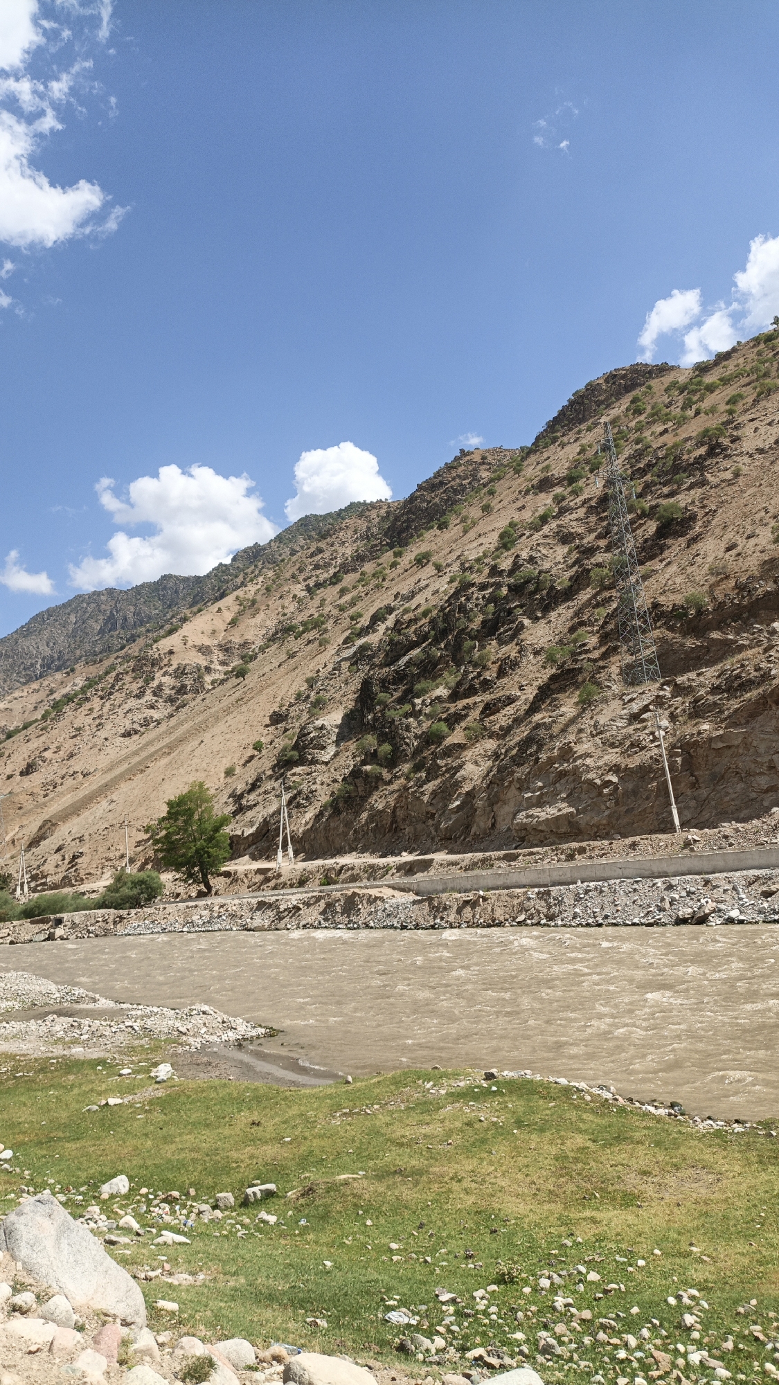A wide, muddy river flows through a valley, with a steep, rocky mountainside rising on the right. The mountainside is sparsely covered with vegetation and features a power line tower. A road runs along the base of the mountain, parallel to the river. The foreground on the left consists of a grassy embankment dotted with rocks. The sky is blue with scattered white clouds. The image appears to be taken in a natural, rugged landscape, likely a rural area outside of a city.