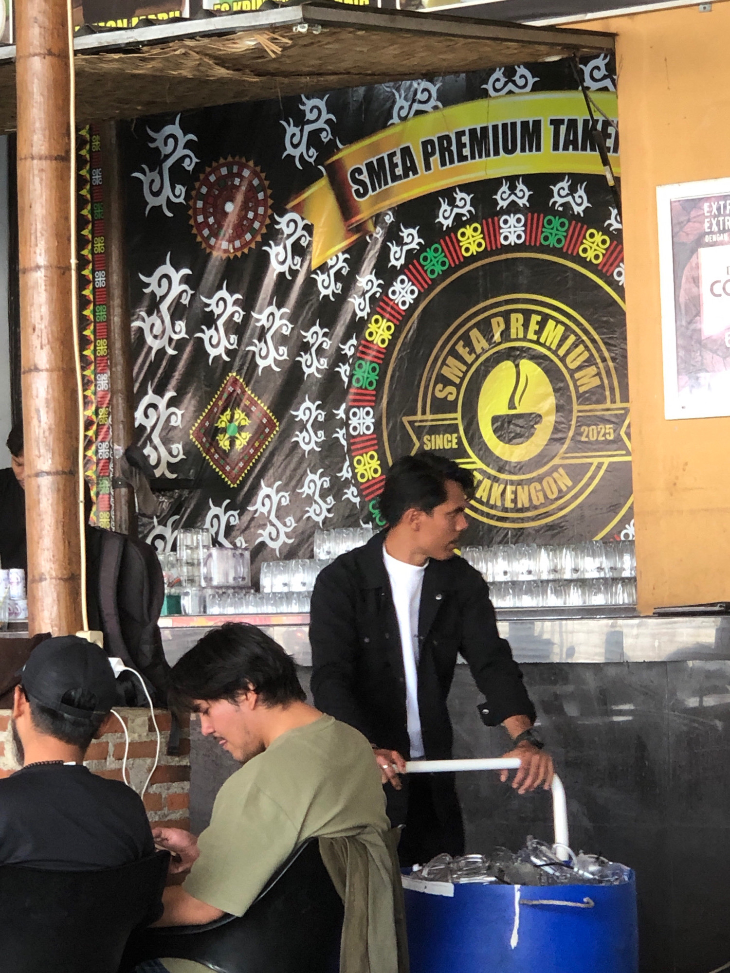 The image captures an outdoor scene at a food or beverage stall in Nunangantara, Indonesia. A man in a black jacket and white t-shirt is standing behind a counter, holding a white pole attached to a blue cooler filled with ice and glasses. He is looking to his right. In the foreground, two men are seated, facing away from the camera. The man on the left wears a black t-shirt and a black baseball cap. The man on the right wears a light green t-shirt. Behind the counter, a banner with traditional Indonesian patterns and the text "SMEA PREMIUM TAKENGON SINCE 2025" is displayed. The environment suggests a casual, open-air setting.