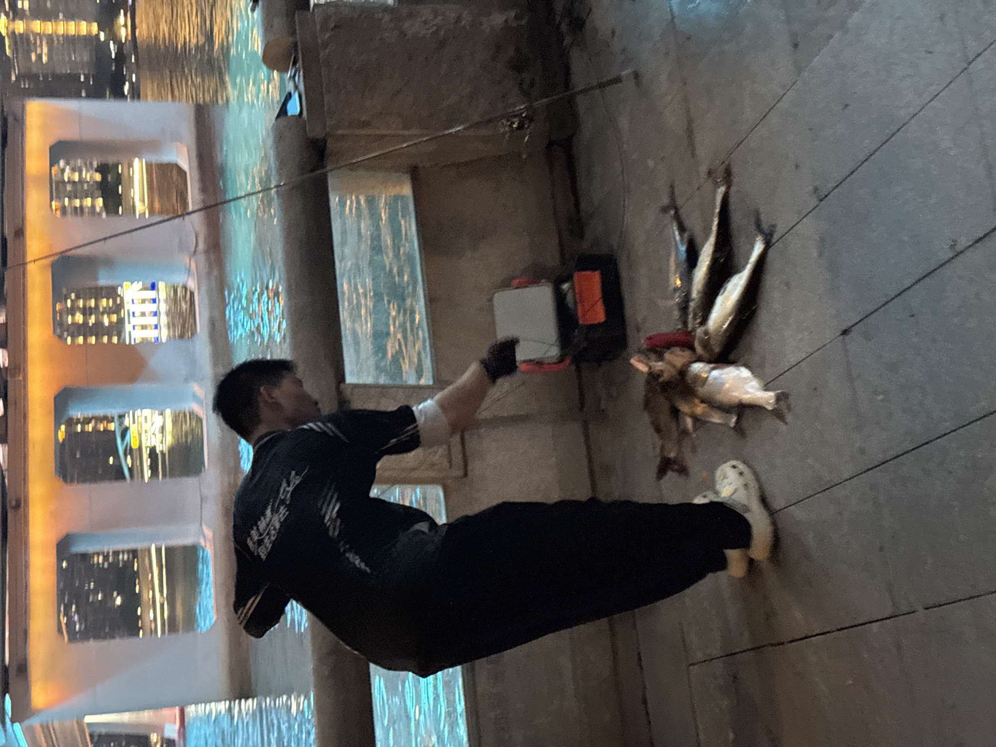 A man is fishing along the river in the Tianhe District of China. He is wearing a black shirt with white stripes on the sleeves, black pants, and white shoes. He has black gloves on both hands. The man is next to the river and a cement bridge. He is holding a fishing rod and has caught a pile of fish that are lying on the ground next to him. There is a fishing box next to the fish. The river reflects the city lights.