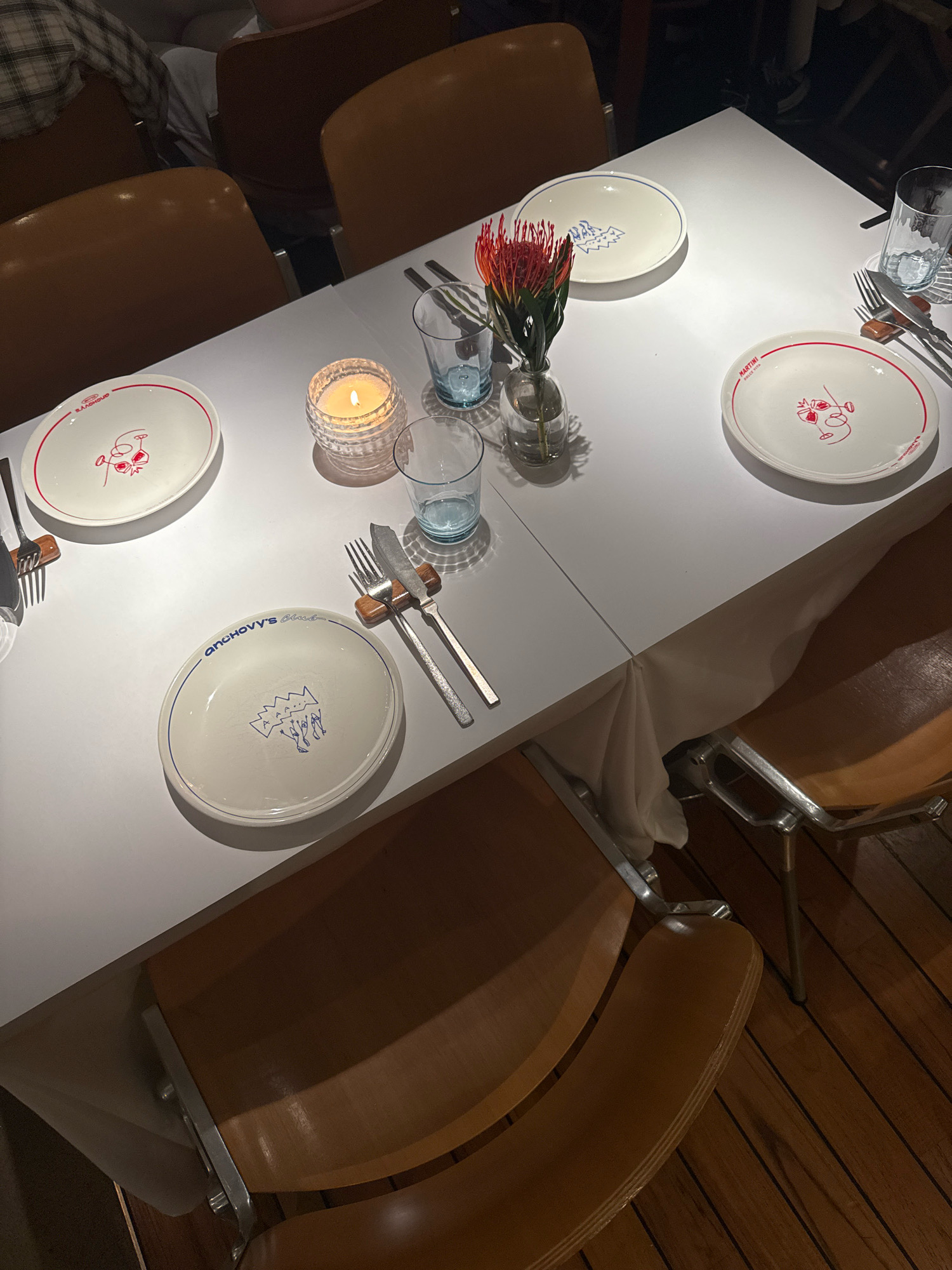 A dimly lit restaurant table setting for a meal is pictured. Four place settings are visible, each with a white plate, silverware, and a blue-tinted glass. Two plates feature red rimmed designs of faces, while the other two have blue drawings of abstract figures and text. A small vase holds a red protea flower in the center of the table. A lit candle in a textured glass container adds to the ambiance. The table is covered with a white tablecloth that drapes onto the floor. The chairs around the table are made of light brown wood with metal legs. The flooring is dark wood. The scene appears to be in a restaurant in Moscow, Russia.