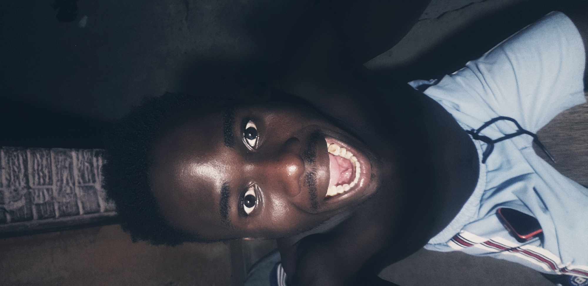 A man is looking up at the camera with his mouth wide open in a laugh. He is bare-chested and wearing light blue shorts with red and white stripes. A dark-colored cell phone is tucked into the pocket of his shorts. The background appears to be a brick wall on the left and a darker, textured surface on the right. The lighting is dim, with highlights on the man's face and shoulders. This image was taken in Narayi, Nigeria.