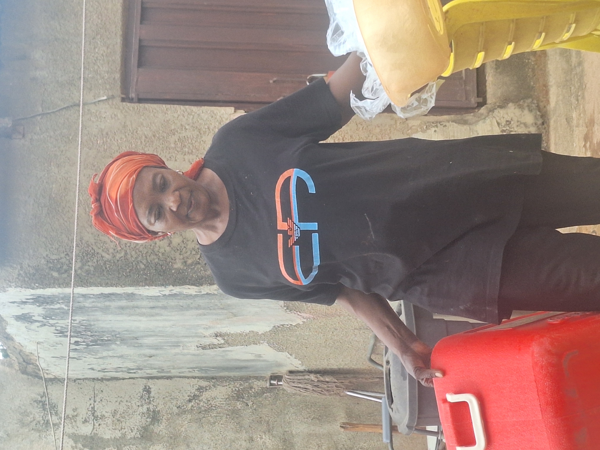 A woman wearing a headscarf and a black t-shirt with a logo is standing outside in Sabon Tasha, Nigeria. She is holding a red cooler with a white handle. To her left, there is a mop. Behind her, there is a yellow chair. The background consists of a concrete wall and a wooden door or window frame. The lighting suggests it is daytime.