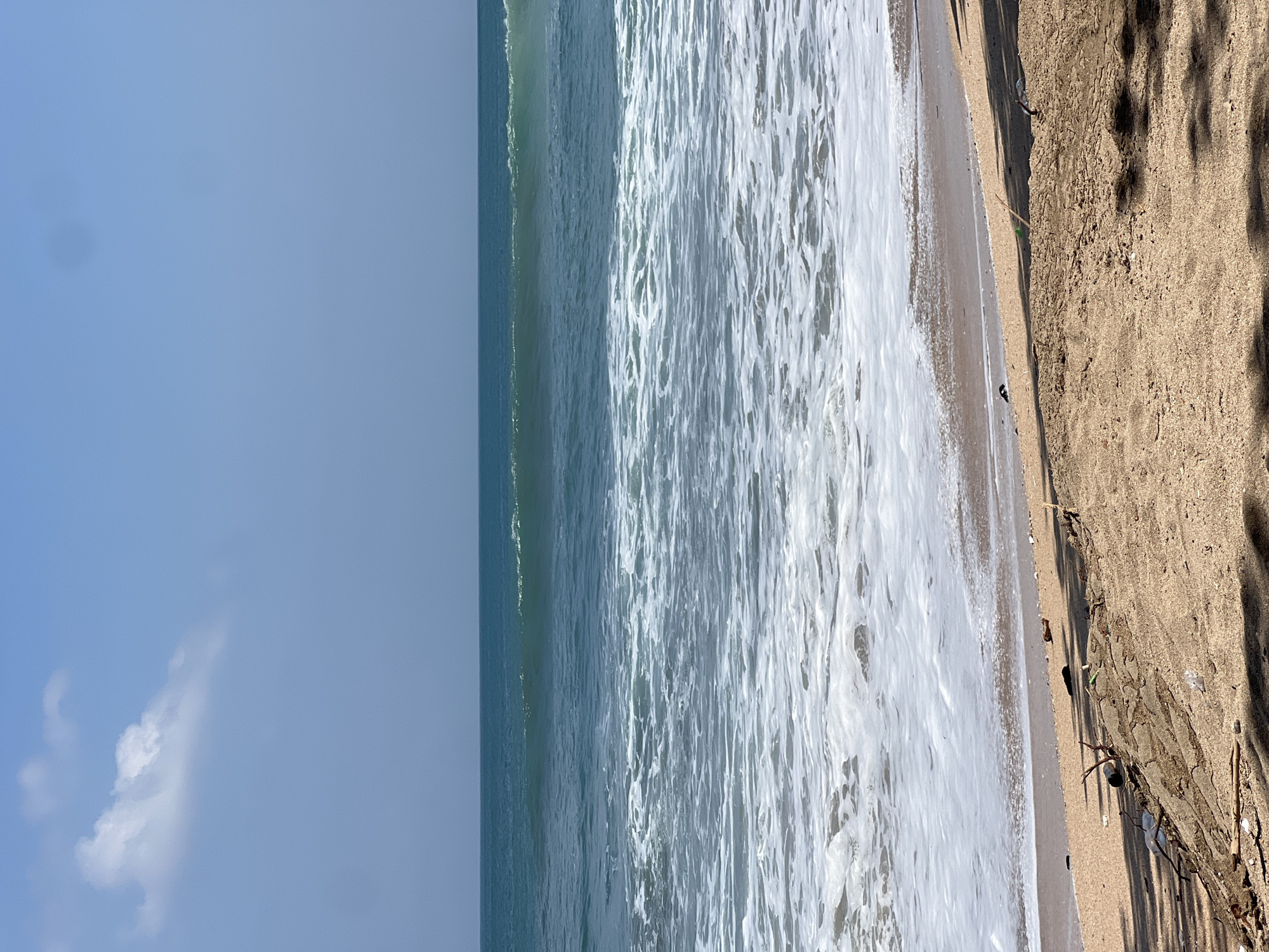 The image depicts a beach scene in Kuta, Indonesia, characterized by a clear blue sky with a few white clouds. The ocean is a vibrant teal, with moderately sized waves breaking and creating white foam that extends towards the shore. The beach itself is sandy and appears to have some debris, including twigs and scattered plastic. The foreground is dominated by the sand, with shadows cast from unseen foliage. The overall impression is a somewhat natural yet affected coastal environment.