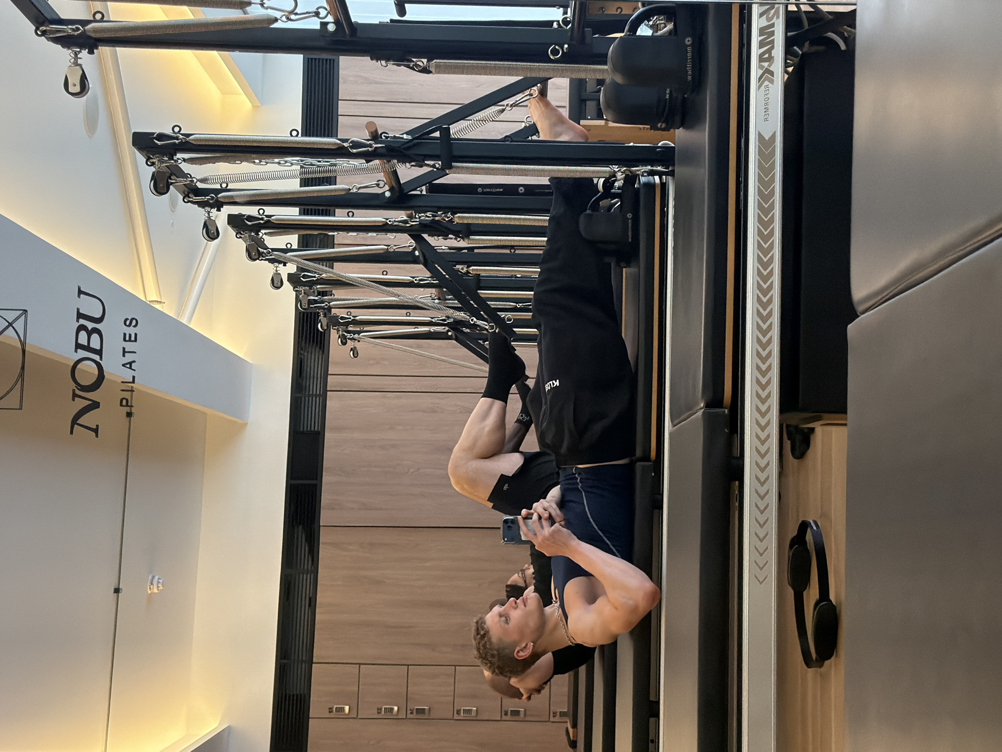 A man is pictured upside down on a Pilates reformer machine. He is fair-skinned with short blonde hair. He is wearing a dark blue tank top and black shorts, and black socks. He is holding a smartphone in his hand and appears to be looking at it.
The Pilates reformer machine is black and metal, with springs and pulleys visible. The word "NOBU PILATES" is printed on a nearby wall. The background consists of light wood paneling, possible lockers or cabinets.
Based on the branding and the Pilates reformer equipment, the location appears to be a Pilates studio, specifically Nobu Pilates, likely in London, United Kingdom.