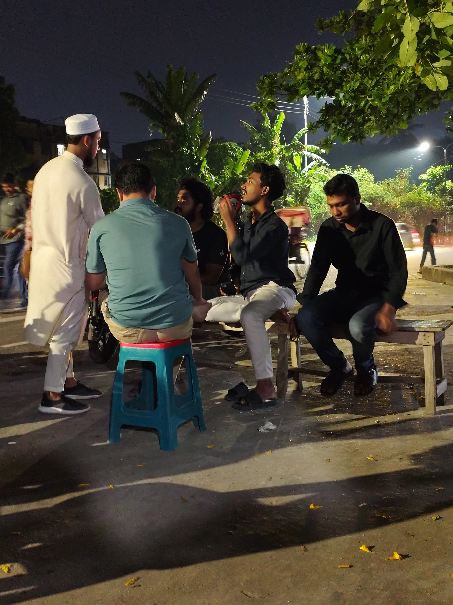 The image depicts a group of men gathered outdoors at night in Dhaka, Bangladesh. Four men are centrally located, one standing and wearing a white tunic and cap, another sitting on a blue plastic stool in a teal shirt, another sitting on a bench in a black shirt drinking from a red cup, and another sitting on the same bench in a black shirt. To the left, two other men are partially visible. In the background, there are banana trees and streetlights. The ground is concrete and littered with yellow leaves. A street is in the background with a passing car.