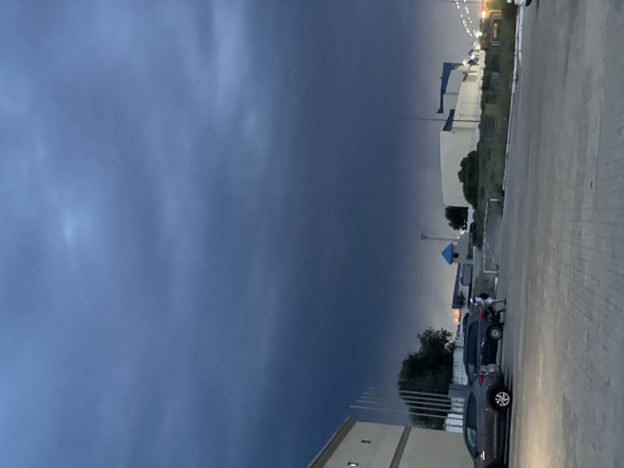 The image captures an outdoor scene at dusk or early evening in Aktau, Kazakhstan. The sky is dominated by dark, heavy clouds, suggesting an impending storm or overcast weather. In the foreground, a paved area shows parked cars, including a dark gray SUV and a dark minivan. A person, possibly a male wearing a white cap and shorts, is seen near the minivan, interacting with its rear. To the left, a portion of a light-colored building with windows is visible. In the midground, there are more buildings, some appearing industrial or commercial, with lights visible in the distance. Vegetation, including trees and bushes, is scattered throughout the scene. The overall atmosphere is somber due to the dark sky.