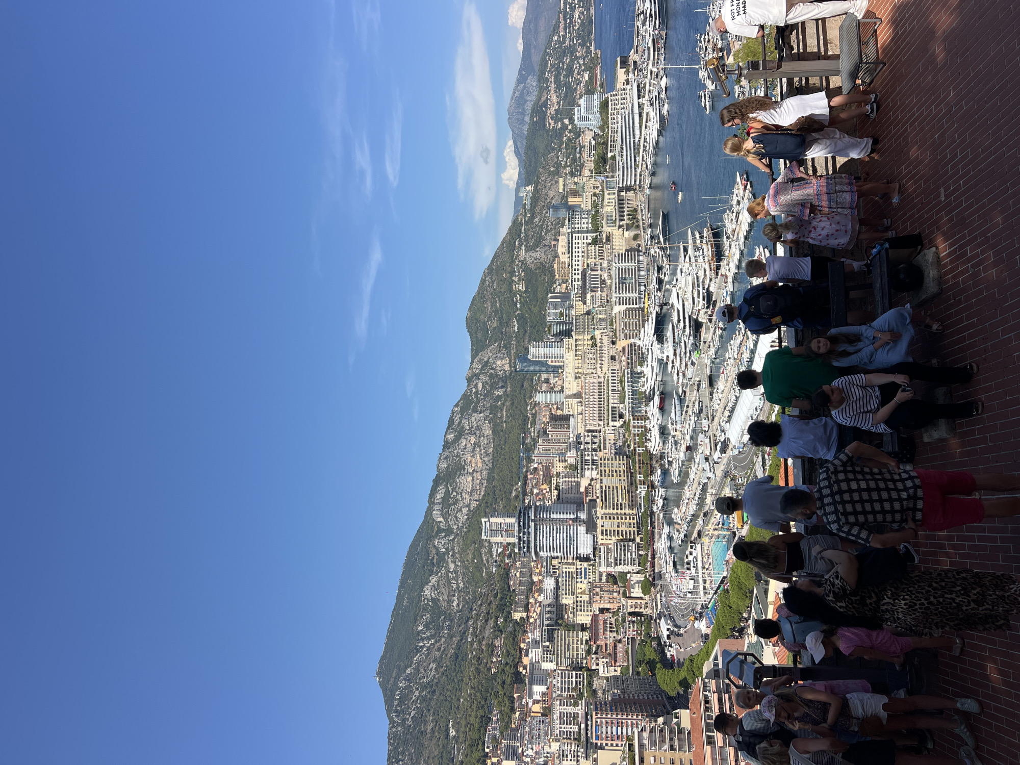 This is a high-angle, wide shot of Monaco, taken on a clear, sunny day. In the foreground, a group of approximately 15-20 people are gathered on a brick-paved overlook, looking out at the city and the harbor. Many are facing away from the camera, observing the view. Some individuals are dressed casually in t-shirts, shorts, and dresses.
The middle ground is dominated by a densely populated city skyline with numerous buildings of varying heights and architectural styles, constructed from materials like concrete, glass, and stucco in colors ranging from white and cream to yellow and pink. A prominent, modern, cylindrical building with a green exterior stands out amongst the others. Below the city, a large harbor is filled with a vast number of docked yachts and smaller boats, their masts creating a dense, white forest. A winding road with visible white lines and a swimming pool are also situated in this area, suggesting a race track or event space.
The background features a steep, green, mountainous terrain covered in trees. The sky is a clear, vibrant blue with a few wispy white clouds. The overall impression is a bustling urban center nestled between mountains and the sea, with a recreational waterfront and a scenic overlook providing a vantage point for visitors.