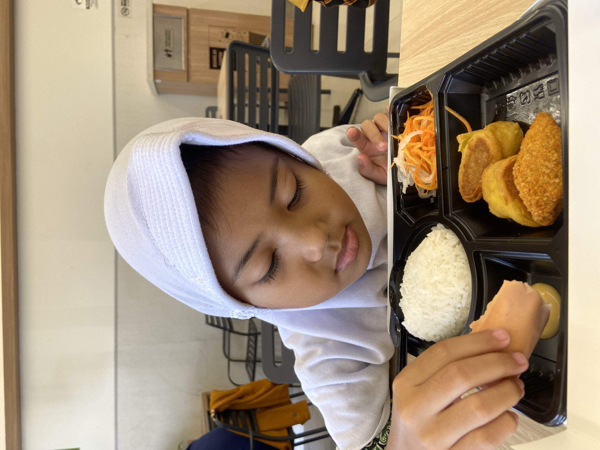 A young person wearing a white head covering sits at a table in what appears to be a cafeteria or food court setting. The individual is holding a hotdog or sausage, seemingly about to eat it. Beside them is a black bento box, portioned into compartments containing a scoop of white rice, a serving of shredded carrots and radish, and several pieces of golden-brown fried food. A small dollop of mustard is visible in the compartment where the hotdog is being held. The seating area features gray chairs with a slatted back design. The backdrop includes neutral-colored walls and suggests an indoor setting with food service facilities, possibly located in Sidoarjo, Indonesia.