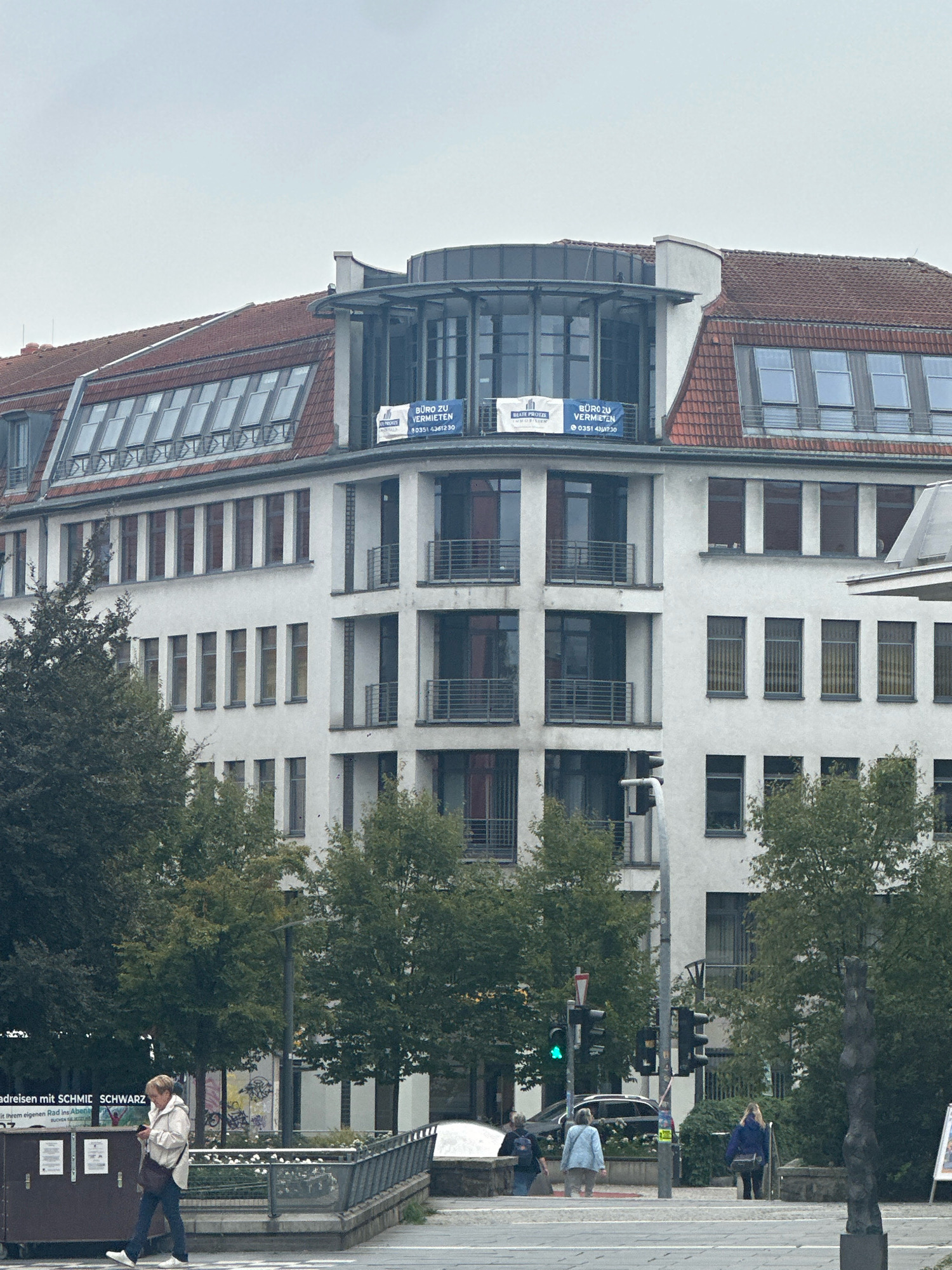 The image depicts a view of a multi-story white building with a red tiled roof, likely in an urban setting. The building features numerous windows, some with awnings and others with balconies. Two blue banners are visible on the upper part of the building, one reading "BÜRO ZU VERMIETEN" (Office for Rent) and a phone number "0351 4361230". Green trees partially obscure the lower floors of the building. In the foreground, a woman in a white jacket and dark pants is walking while looking at her phone. To the right, there are traffic lights, one of which is green. Several other individuals are visible in the distance, some walking and others standing. The overall environment appears to be a street or pedestrian area with some urban landscaping. The image is taken from a slight distance, suggesting a street-level perspective.