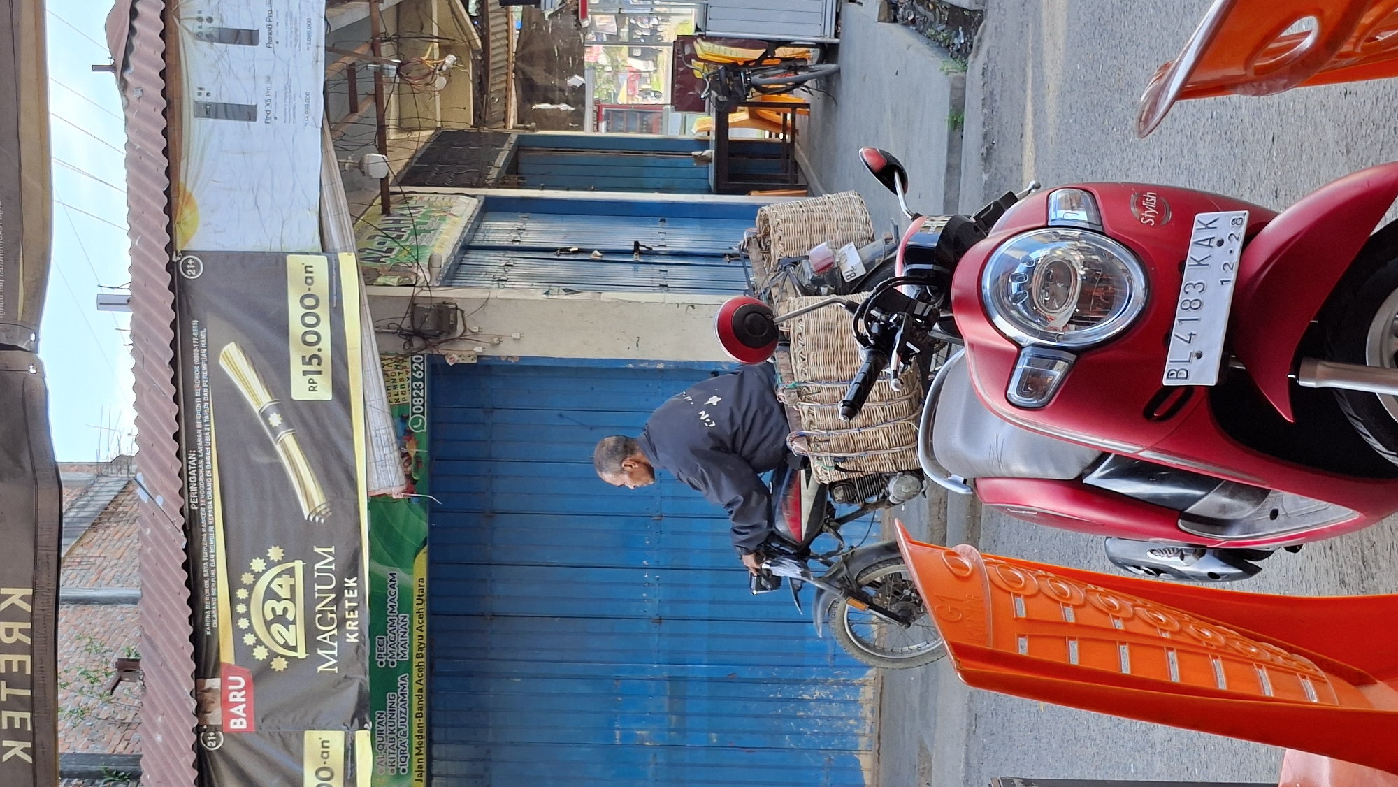 This image captures a street scene in Keudebayu, Indonesia, likely during the daytime given the bright sunlight casting shadows. The central focus is a red scooter parked on a paved surface, adorned with two woven baskets. A man, seemingly a vendor, is visible in the background, bent over near the scooter, possibly engaged in loading or unloading goods.
The scooter has a license plate reading "BL 4183 KAK 12-28" and the word "Stylish" is emblazoned on its side. In front of the scooter, an orange object, possibly a protective cover or accessory, is prominent. The background reveals a row of blue storefronts with closed shutters, suggesting it might be a quieter part of the day or a business that operates differently. A corrugated metal roof is visible on the left, indicating a typical building style in the area.
A prominent banner on the left side of the image advertises "MAGNUM KRETEK" cigarettes, with a price of "RP 15.000-an" and a "BARU" (new) indication. It also includes a warning about smoking and a contact number: "0823 620". Another banner above it displays images of what appear to be mobile phones, with text including "Find X5 Pro". The overall impression is that of a commercial street in a moderately populated area, with the scooter likely used for delivery or transport of goods, possibly related to the cigarette or other local businesses. The scene feels ordinary and everyday, without any discernible strong emotions or unusual events.