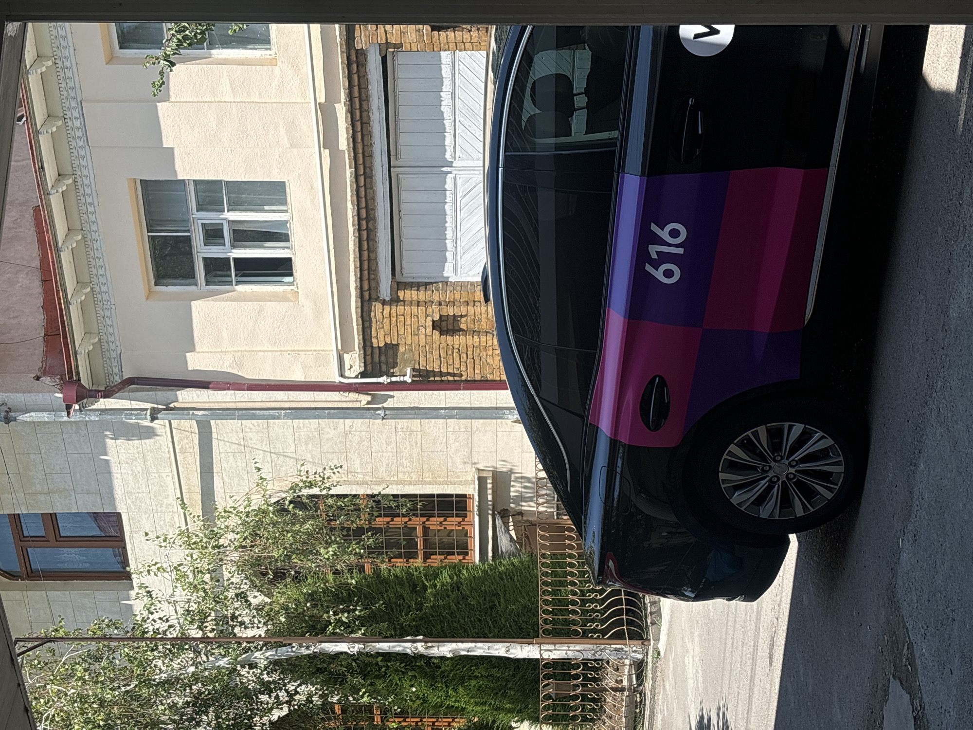 The image captures a sunny daytime scene in the city of Tashkent, Uzbekistan, focusing on the side of a black car parked on an asphalt surface. The car features a distinctive design with colored squares in shades of purple and pink on its rear doors, along with the number "616" in white. A white circular logo with a dark letter inside is also visible on the rear quarter panel. The background reveals a building with a cream-colored facade and brickwork, featuring arched windows with wooden frames and rectangular windows. Green trees and a metal fence are also present in the environment. The scene is viewed from an angle that suggests the photograph was taken from a covered or shaded area.