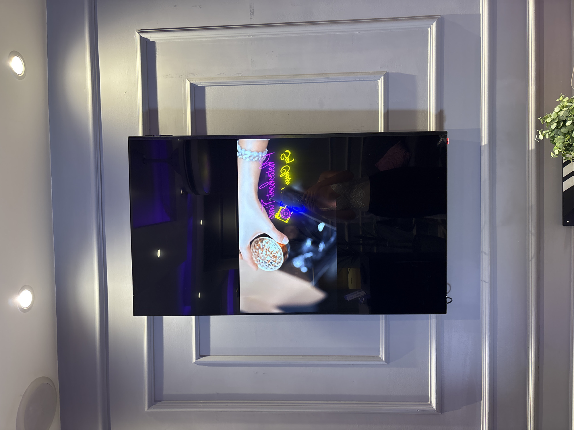 In Sweileh, Jordan, a vertical, black, rectangular screen is mounted on a light purple, paneled wall. The screen displays a video feed showing a person's hand holding a clear glass filled with brown material. Below the hand, a neon sign with yellow and pink lettering is visible, with a blue neon camera icon to the left. A reflection on the screen shows a person wearing a sparkly silver top and black pants, holding a phone. The wall features decorative molding, and a white circular speaker is installed on the ceiling above. The lighting on the wall is soft, with a focused spotlight from the ceiling casting a shadow on the molding.