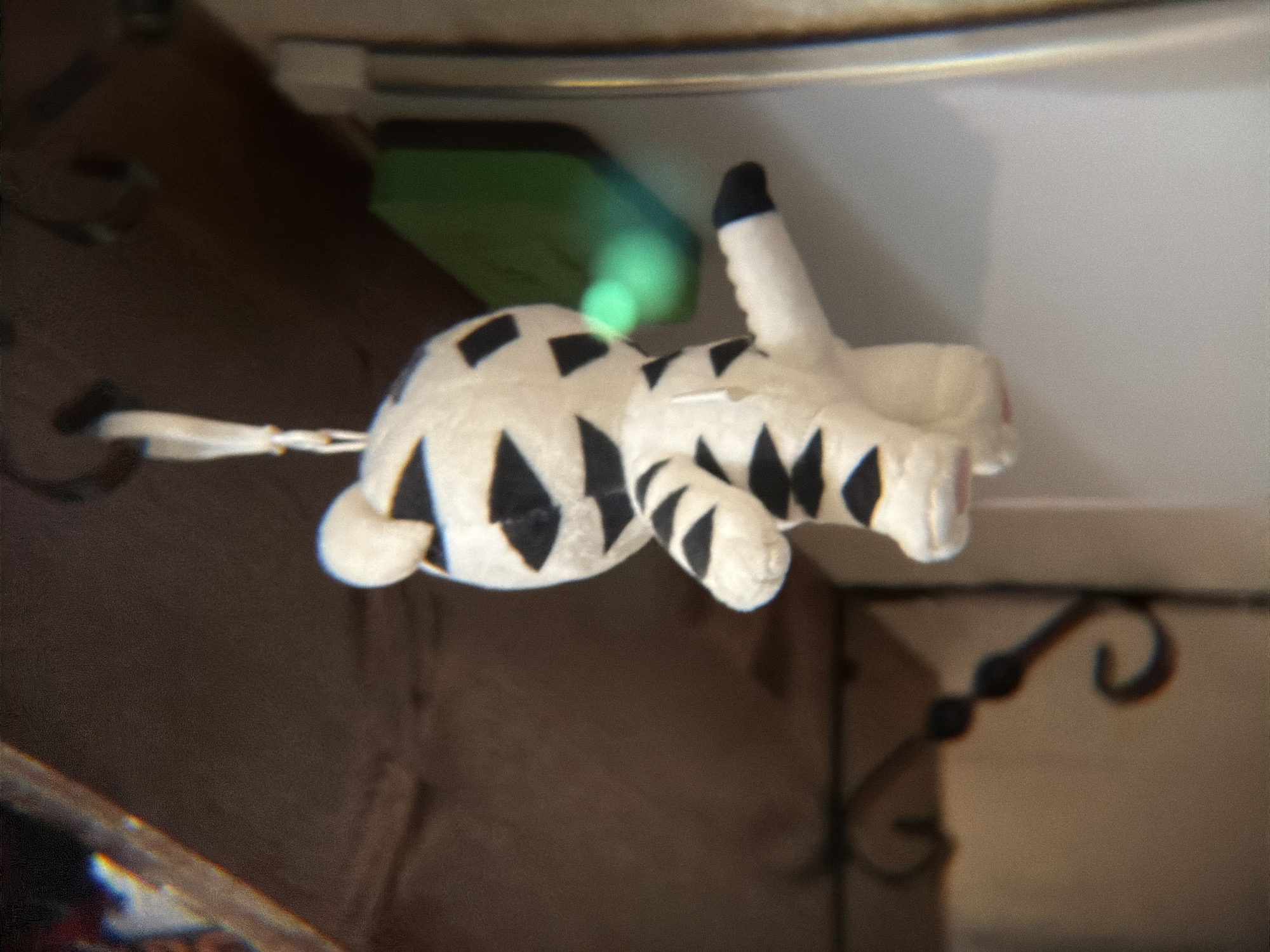 A white plush animal with black markings resembling a tiger is the central object, appearing to float mid-air within the scene. It has a long tail and a raised front leg, suggestive of a walking pose. Background details include a brown wooden structure to the left and lower part of the image, a white surface with a metallic edge above, and an indiscernible green object behind the plush animal. A wrought iron element is also visible in the lower right area of the image. The location is not discernable from the given image.