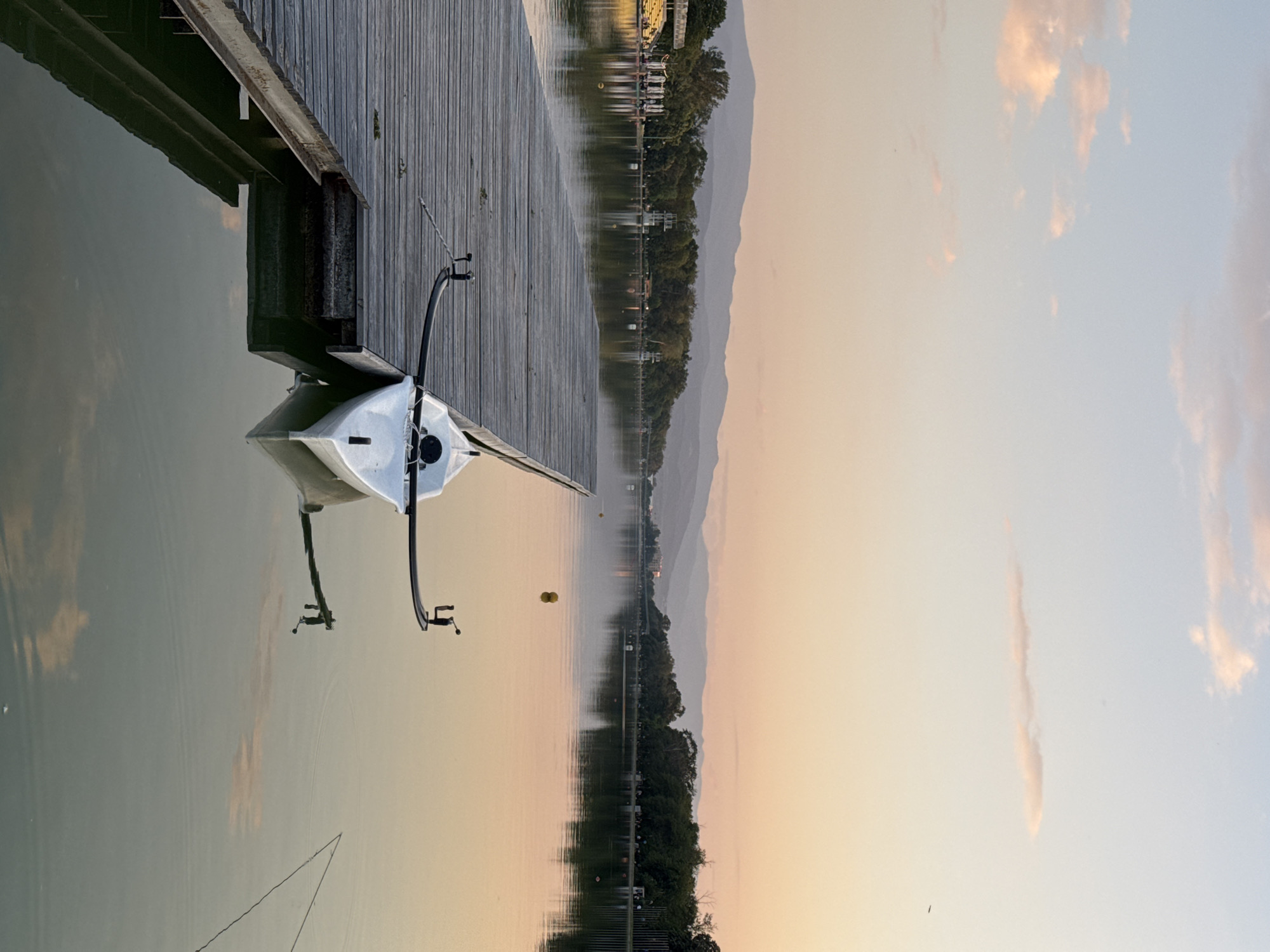 The image depicts a tranquil scene at the Plovdiv Rowing Canal in Bulgaria at dawn or dusk. A wooden dock extends into the calm water, where a white rowing boat is moored. The sky displays a soft gradient of colors, suggesting either sunrise or sunset. The water reflects the colors of the sky, the surrounding greenery, and distant mountains. The scene is still and quiet, with no visible people.