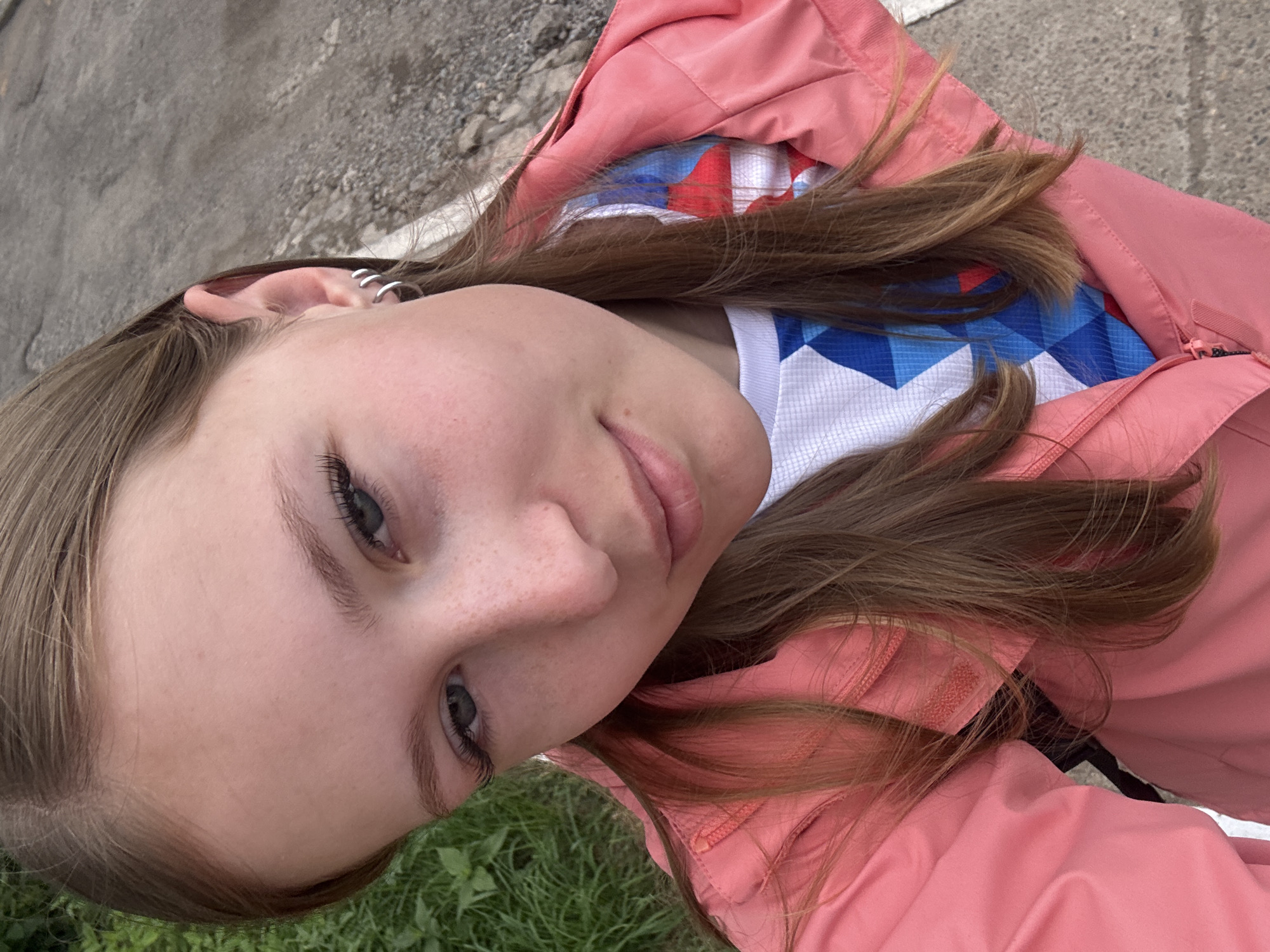 A young girl with light brown hair is taking a selfie outdoors. She is wearing a pink jacket over a white t-shirt with a blue and red geometric pattern. Her left ear, adorned with two silver hoop earrings, is visible. The background shows a paved surface and some green grass.