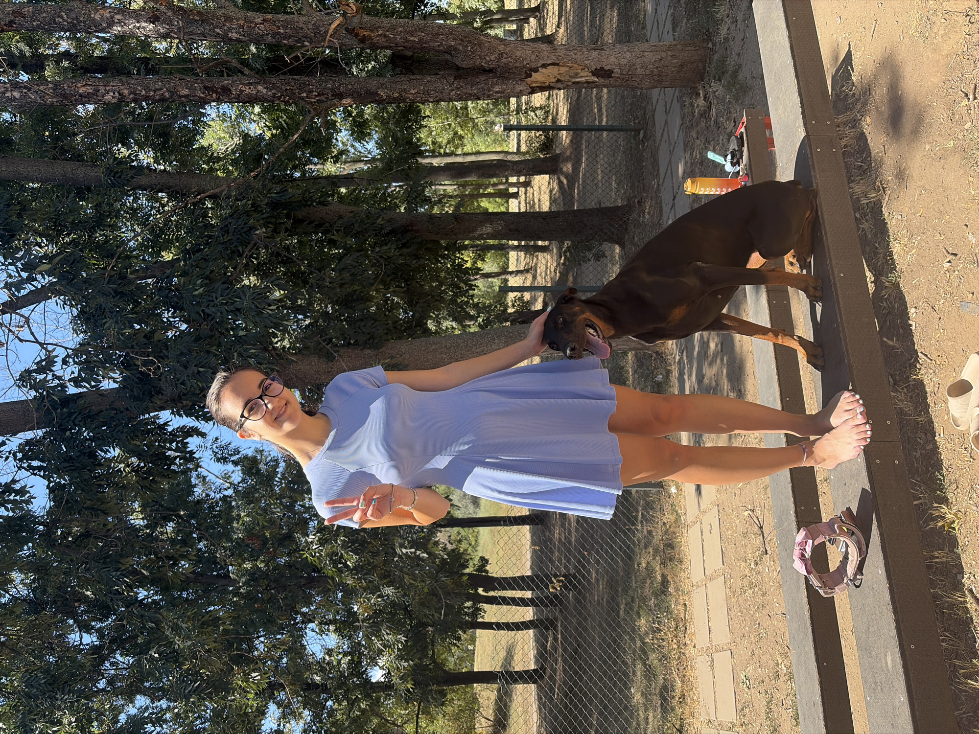 A young woman with glasses and a blue dress is standing in a park, posing for a picture with her dog. The dog, a dark brown Doberman, is sitting next to her. The woman is holding up two fingers in a peace sign. The park has trees, a chain-link fence, and a paved area. There is a dog collar on a wooden bench in the foreground. The scene is well-lit by sunlight. This park is likely located in Sofia, Bulgaria.