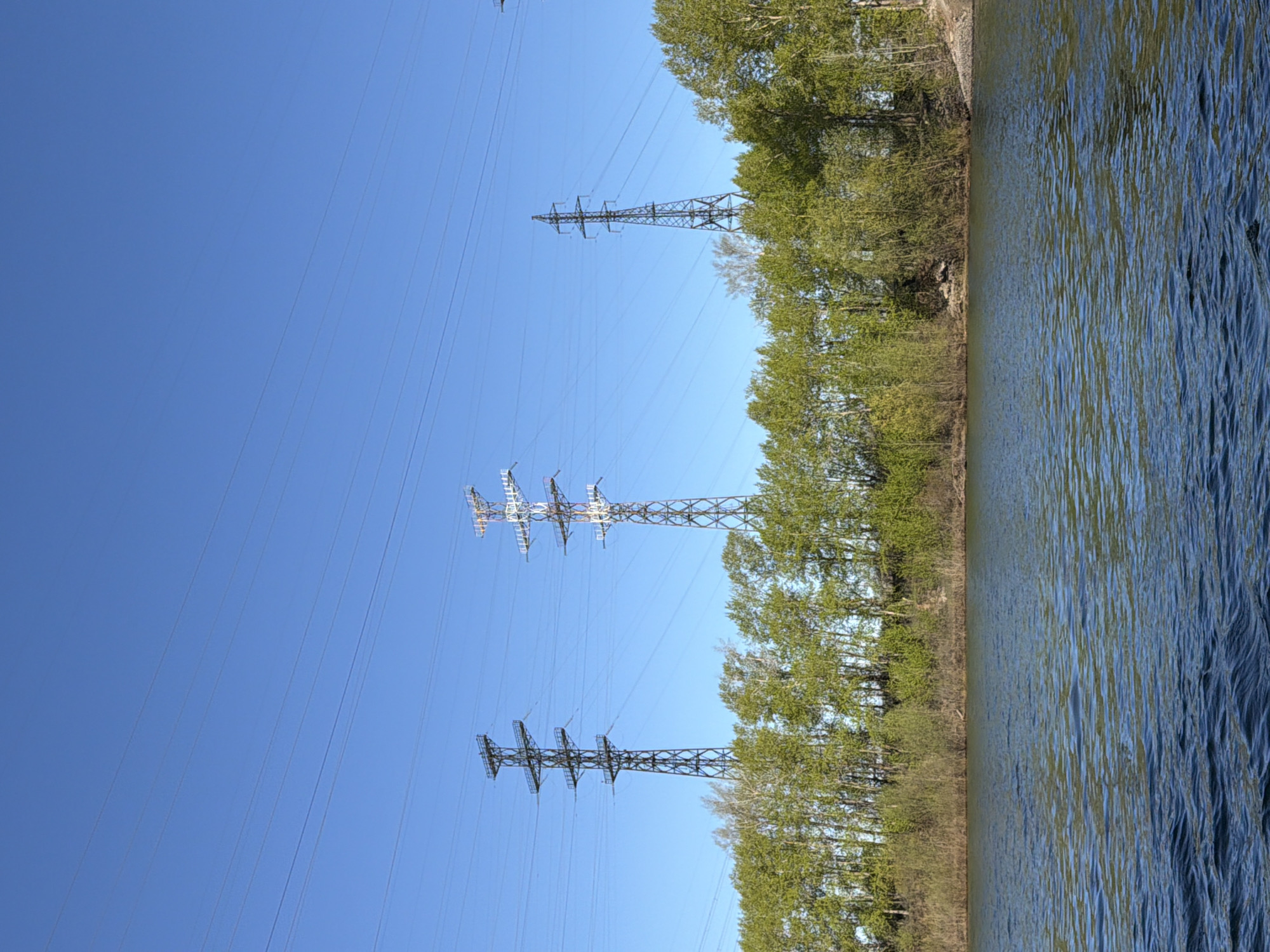 The image shows a landscape in Chaikovsky, Russia, featuring a body of water (likely a river or lake) on the right side, bordered by trees along the bank. Power lines and pylons are visible against a clear blue sky on the left. There are no people visible in the image.