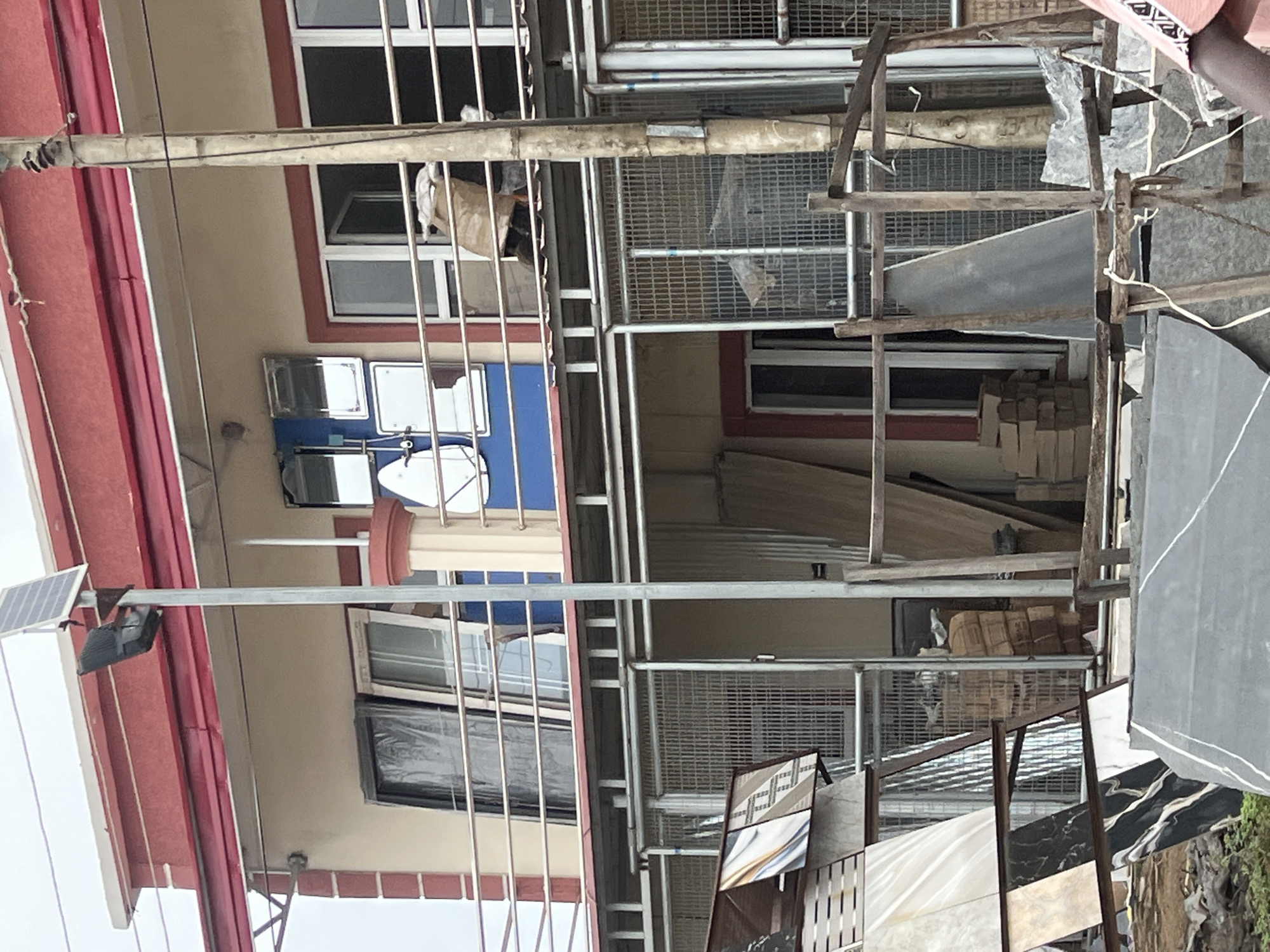 The image shows a close-up view of a building exterior under construction or renovation in Jalingo, Nigeria. A solar panel is mounted on the side of a red-trimmed building, connected to a black light fixture. Below this, there are several windows, some with plastic coverings. A satellite dish is visible on the second floor. The building appears to be multi-story, with metal scaffolding and mesh erected in front of it.
In the foreground, there are various building materials, including large sheets of what appear to be marble or granite, stacked wooden planks, and bundles of tiles or packaged materials. Some of these materials are leaning against the scaffolding or placed on the ground. The overall impression is one of ongoing construction work.
The lighting suggests it is daytime, with bright overcast conditions. There are no visible people in the frame, but a light-colored bag or cloth is seen hanging in one of the windows on the upper floor. The scene is typical of an urban or semi-urban construction site in Nigeria. No text is legible in the image.